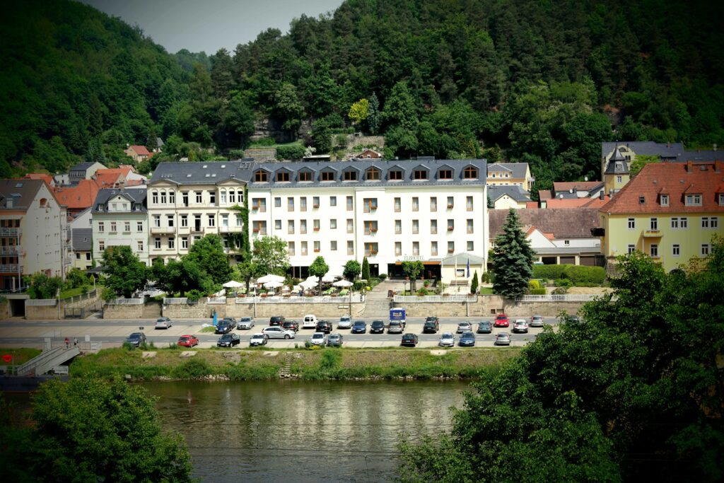 Ein modernes Hotelgebäude mit mehreren Fenstern und einem klaren weißen Design, umgeben von Bäumen und einer Straße mit parkenden Autos.