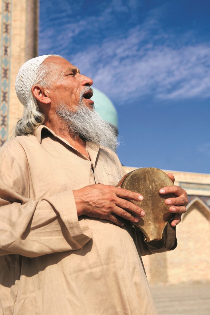 Ein älterer Mann mit grauem Bart und traditioneller Kleidung hält ein Musikinstrument in der Hand und singt im Freien.