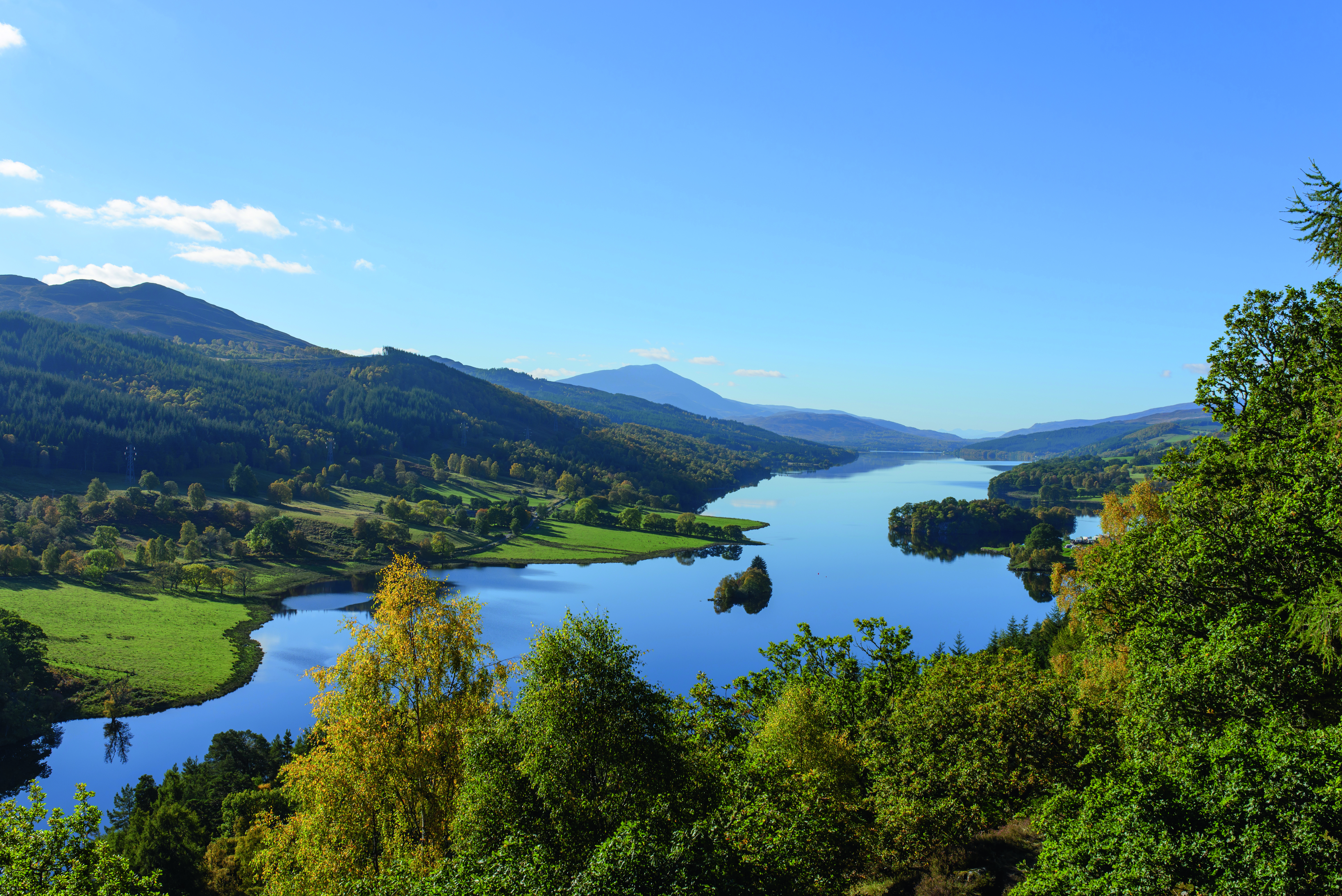 Landschaft mit einem ruhigen See, umgeben von grünen Wäldern und Bergen unter klarem blauen Himmel.