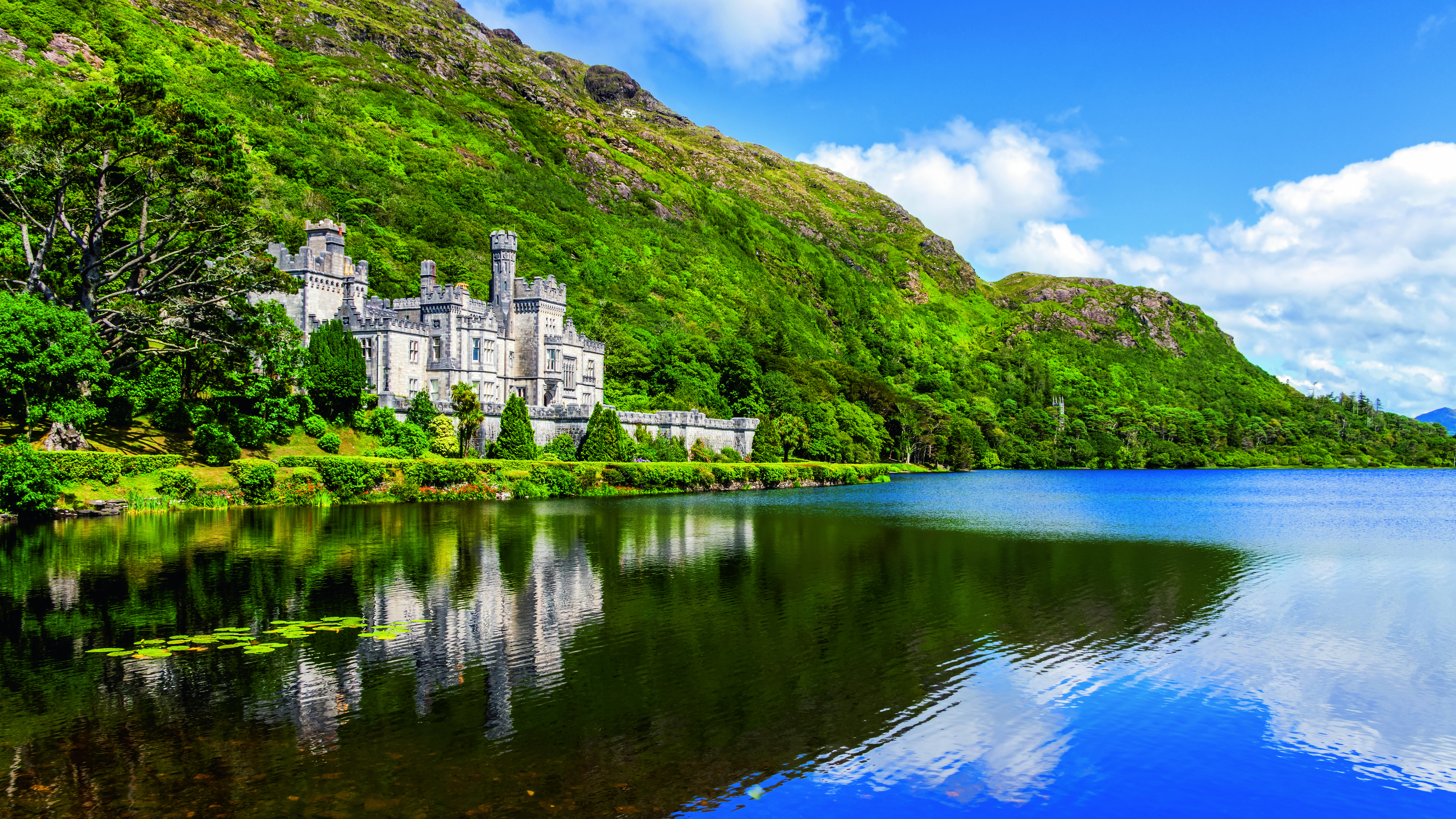 Kylemore Abbey, ein historisches Schloss in Irland, spiegelt sich im ruhigen Wasser eines Sees, umgeben von grünen Hügeln.