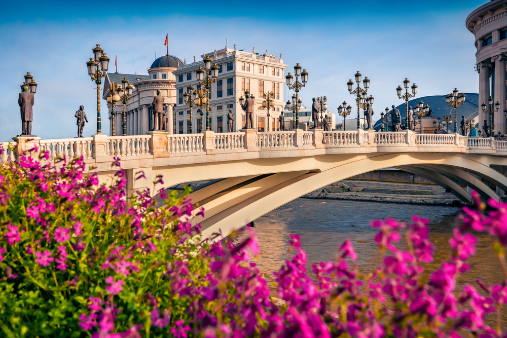 Brücke in Skopje mit Statuen und Laternen, umgeben von blühenden Pflanzen.