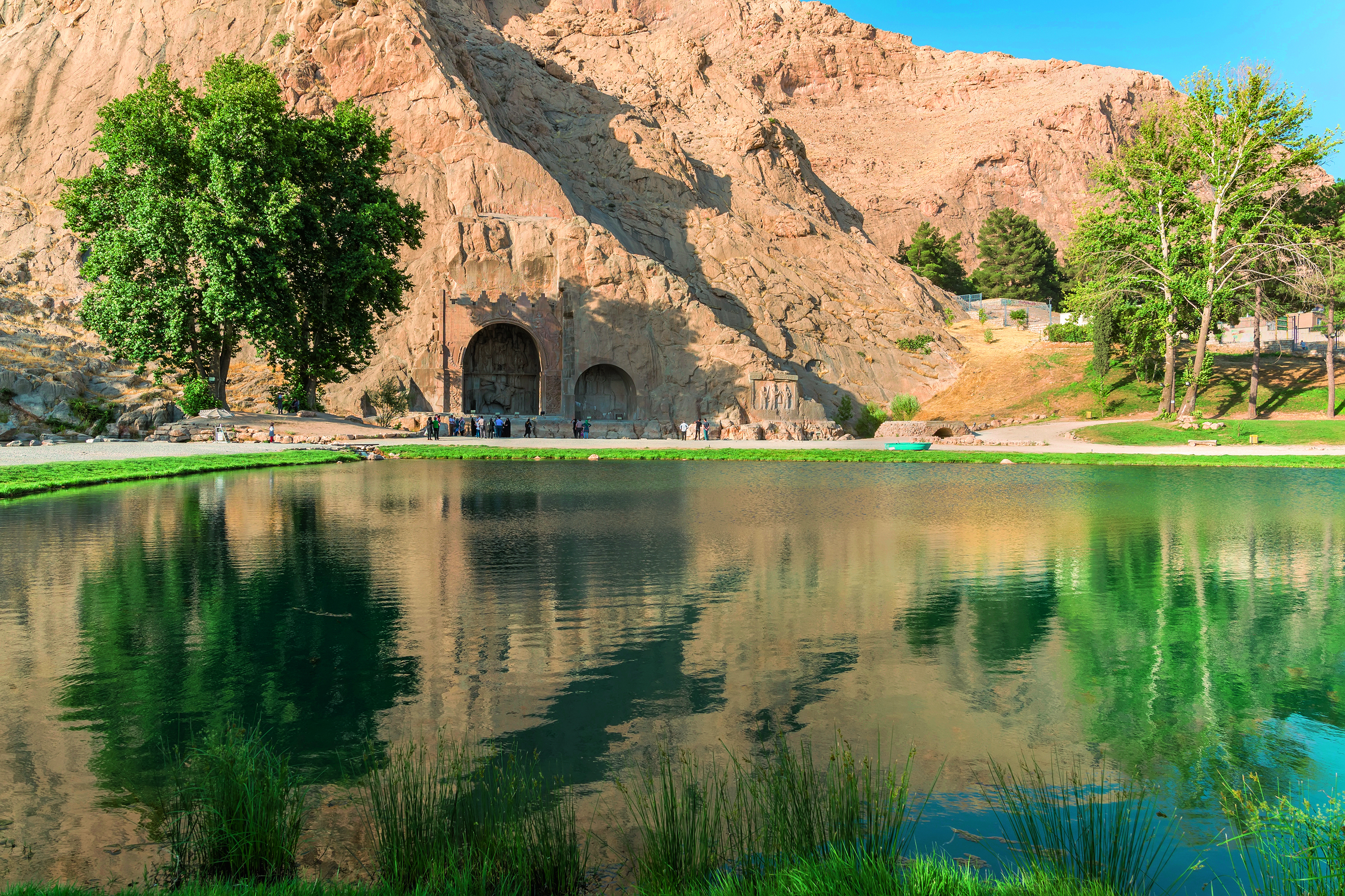 Reflektierendes Wasser vor den Felsengräbern von Taq-e Bostan in Kermanshah, umgeben von Bäumen und Bergen.