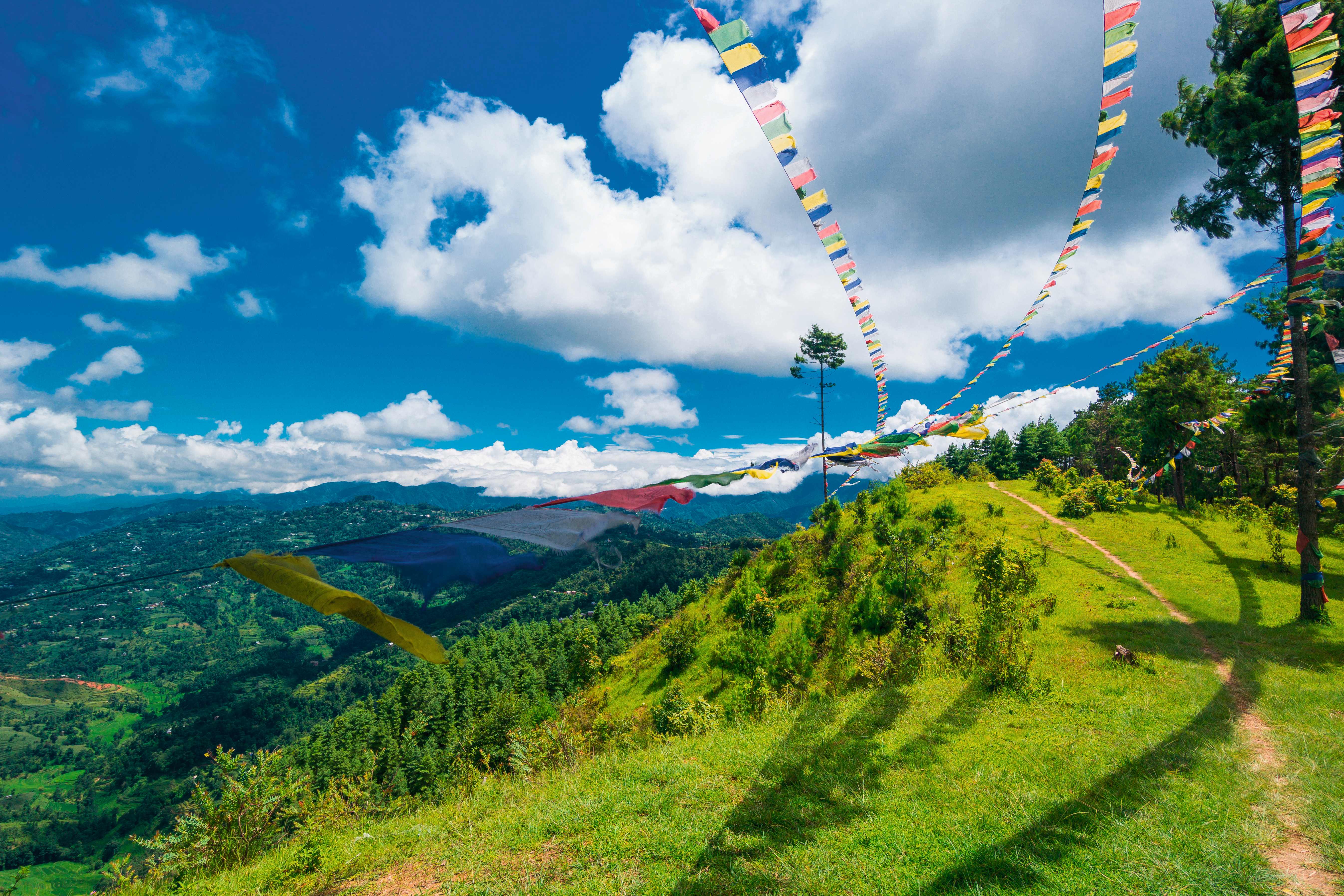 Bunte Gebetsfahnen wehen im Wind über einer grünen Hügellandschaft in Nepal unter einem blauen Himmel.