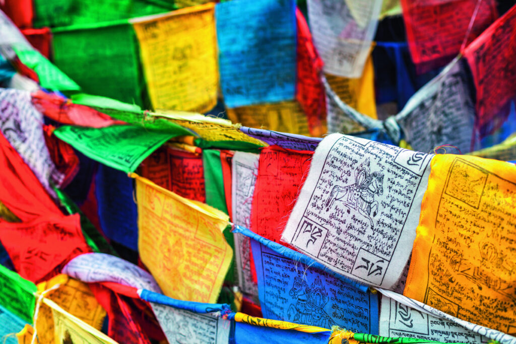 Bunte Gebetsfahnen in verschiedenen Farben, die in der Luft wehen und mit tibetischen Schriftzeichen bedruckt sind.