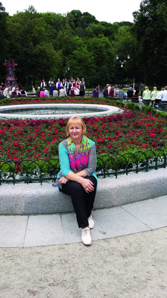 Frau mit buntem Schal sitzt auf einer Steinbank vor einem Blumenbeet in einem Park.