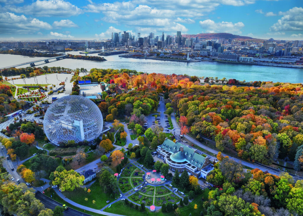 Luftaufnahme eines Parks mit buntem Laub, einem runden Glaskonstruktion und einer Stadtansicht im Hintergrund.