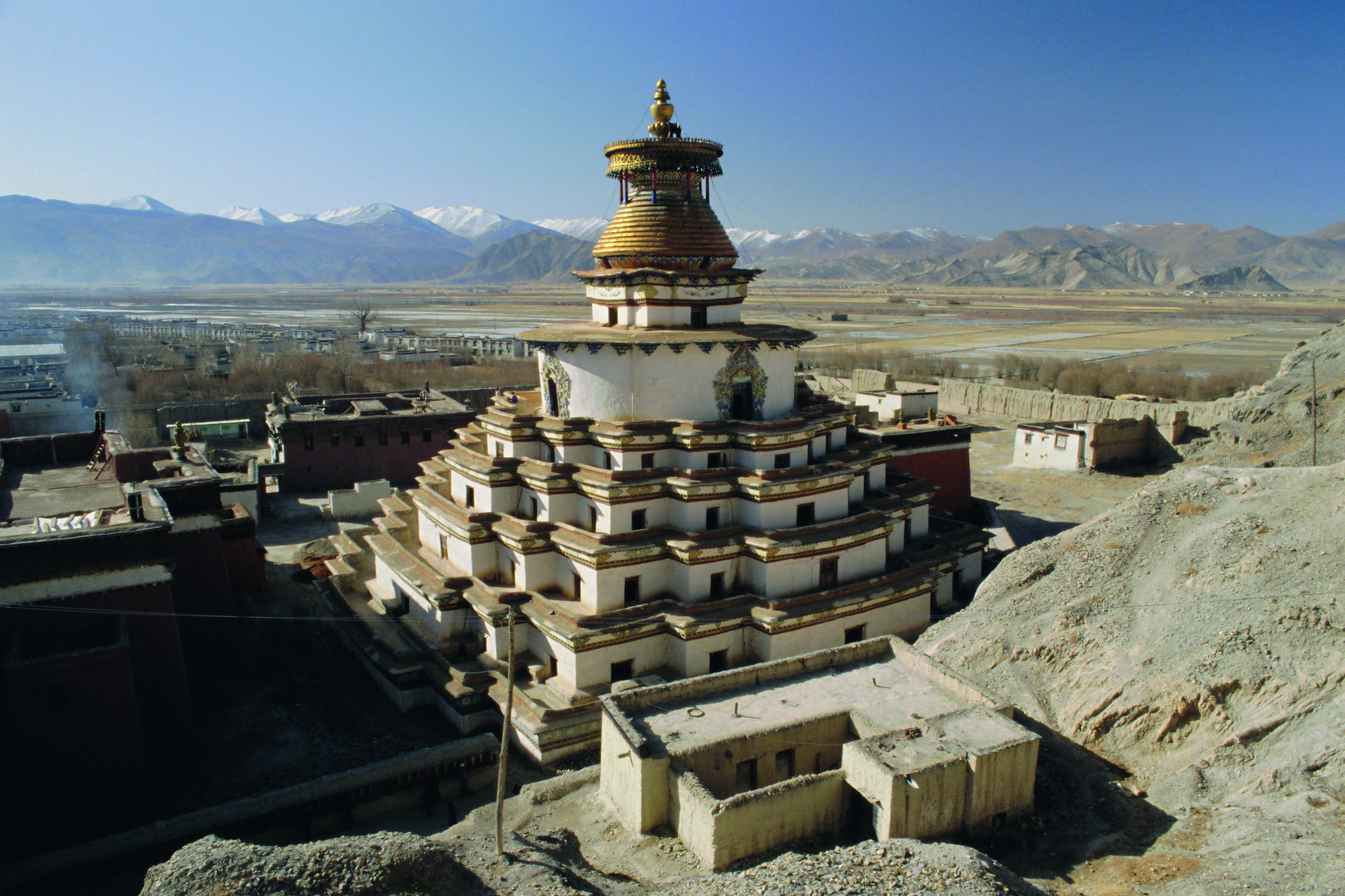 Stupa mit goldener Kuppel und mehreren Ebenen, umgeben von Bergen und einer klaren blauen Himmel.