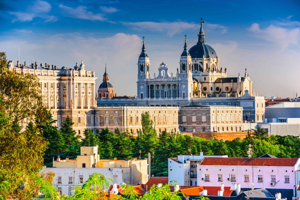 Kathedrale Santa María Real in Madrid, umgeben von Bäumen und Gebäuden, unter blauem Himmel.