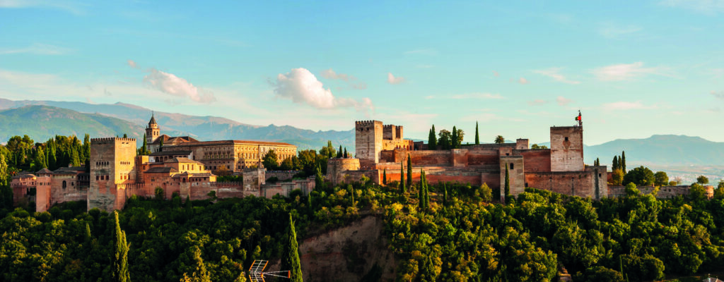 Panoramaansicht der Alhambra in Granada, umgeben von Bäumen und Bergen unter blauem Himmel.