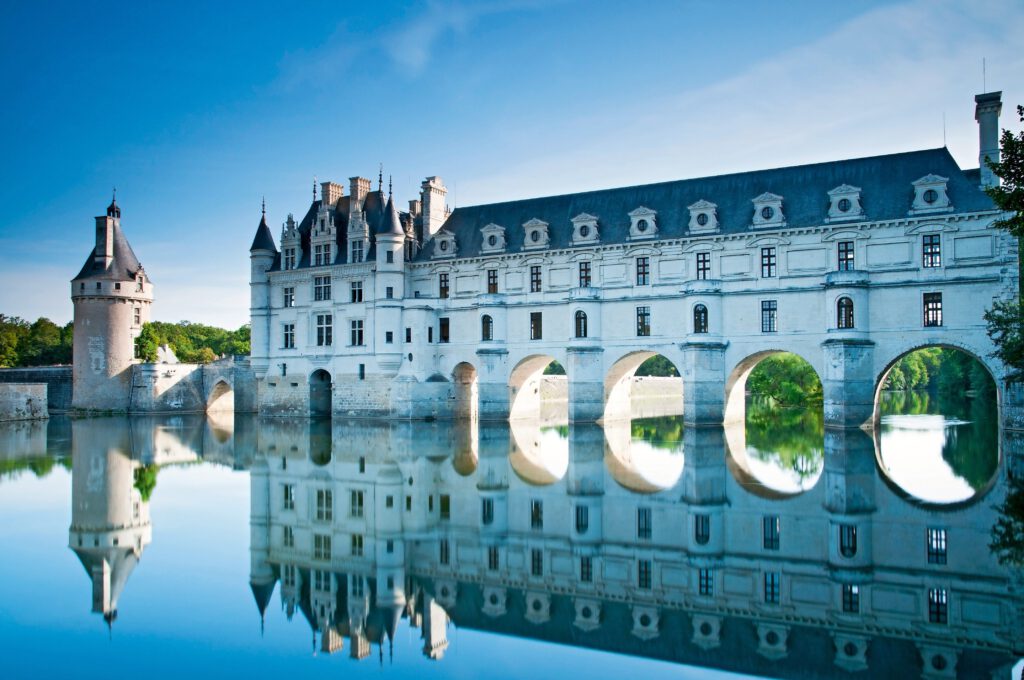 Schloss Chenonceau mit reflektierenden Bögen im Wasser und klarem Himmel im Hintergrund.