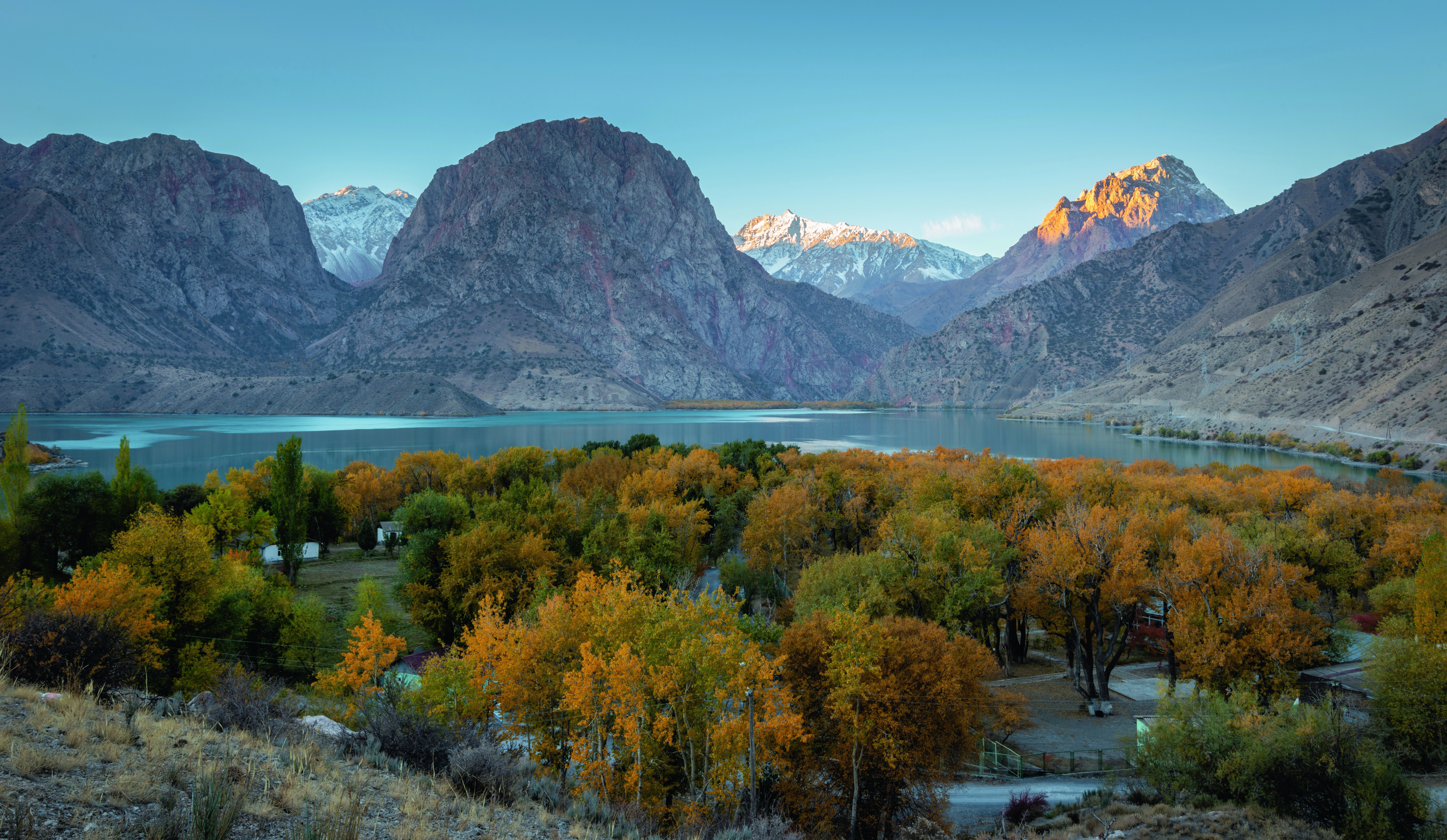 Berglandschaft mit schneebedeckten Gipfeln und einem ruhigen See, umgeben von herbstlichen Bäumen in leuchtenden Farben.