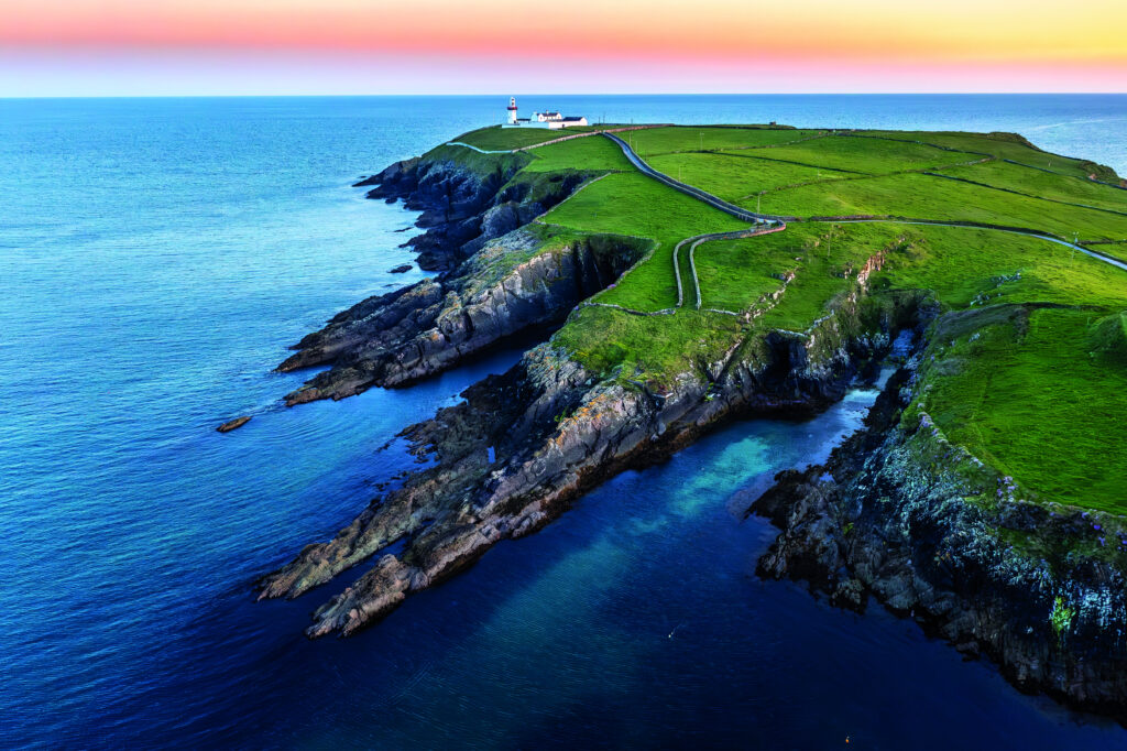 Landschaft mit Küstenlinie, Klippen und einem Leuchtturm auf einer grünen Halbinsel am Meer.