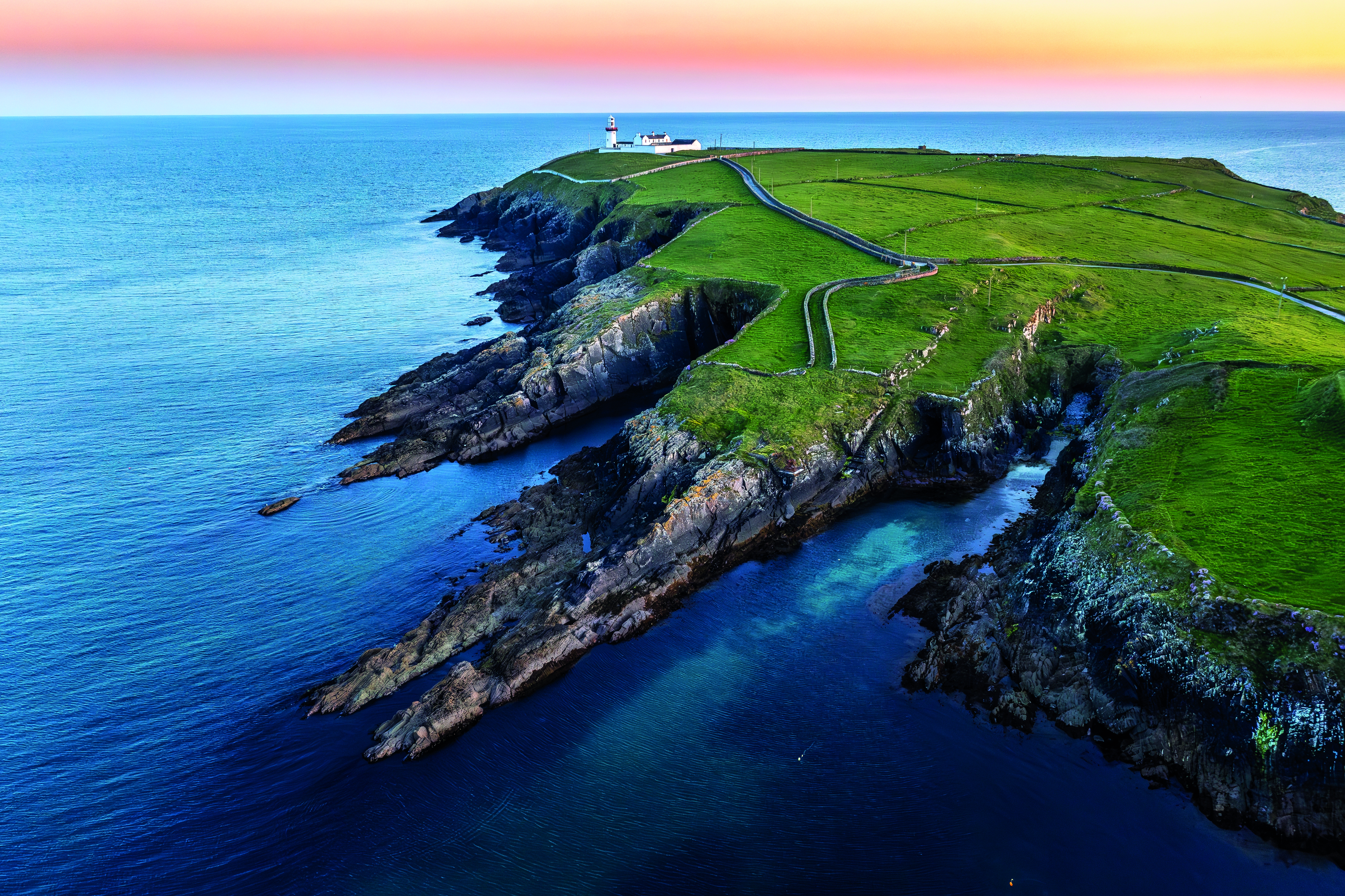 Landschaft mit Küstenlinie, Klippen und einem Leuchtturm auf einer grünen Halbinsel am Meer.
