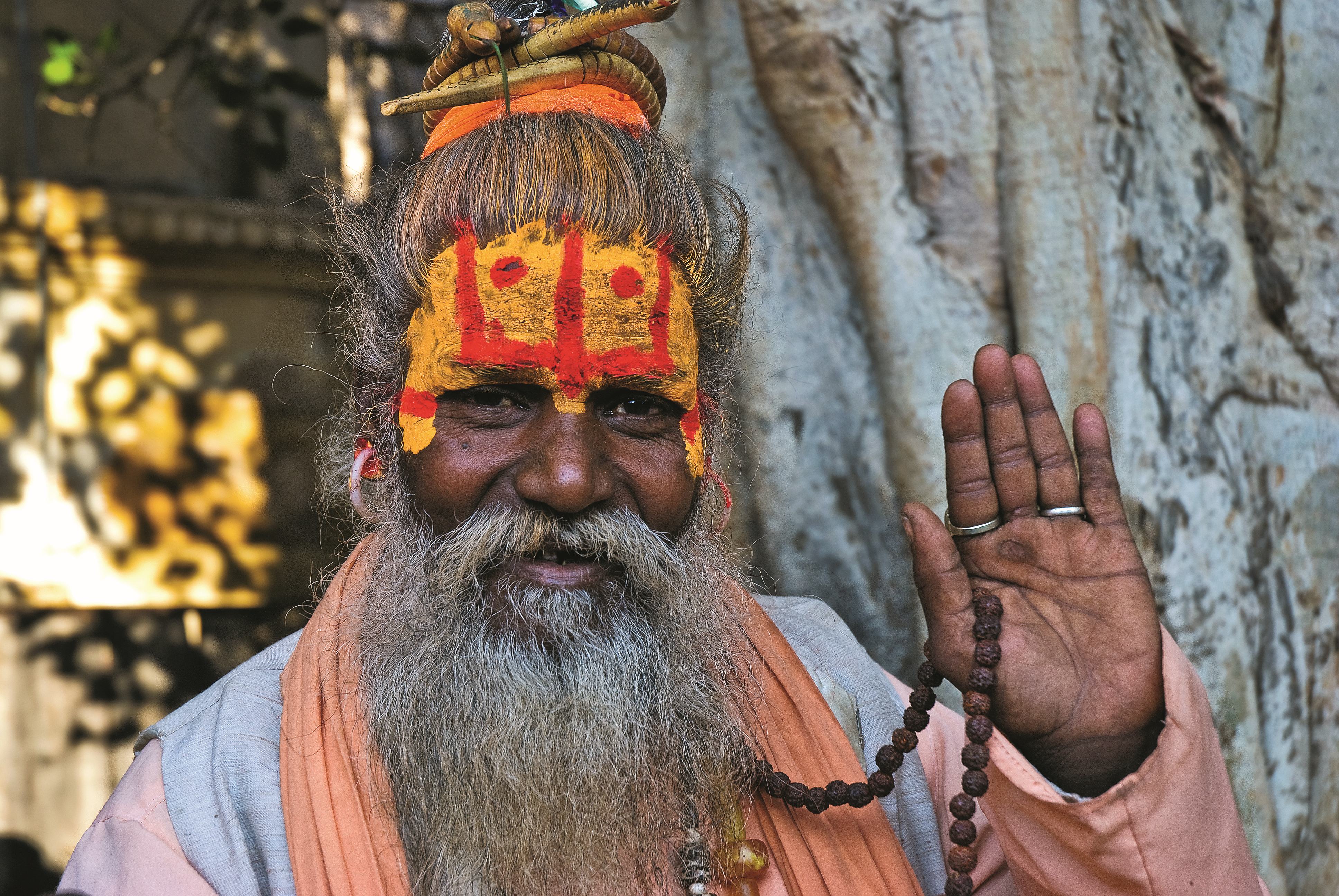 Ein Mann mit orangefarbener Kleidung und bemaltem Gesicht, der mit einer Hand winkt und eine Gebetskette hält.