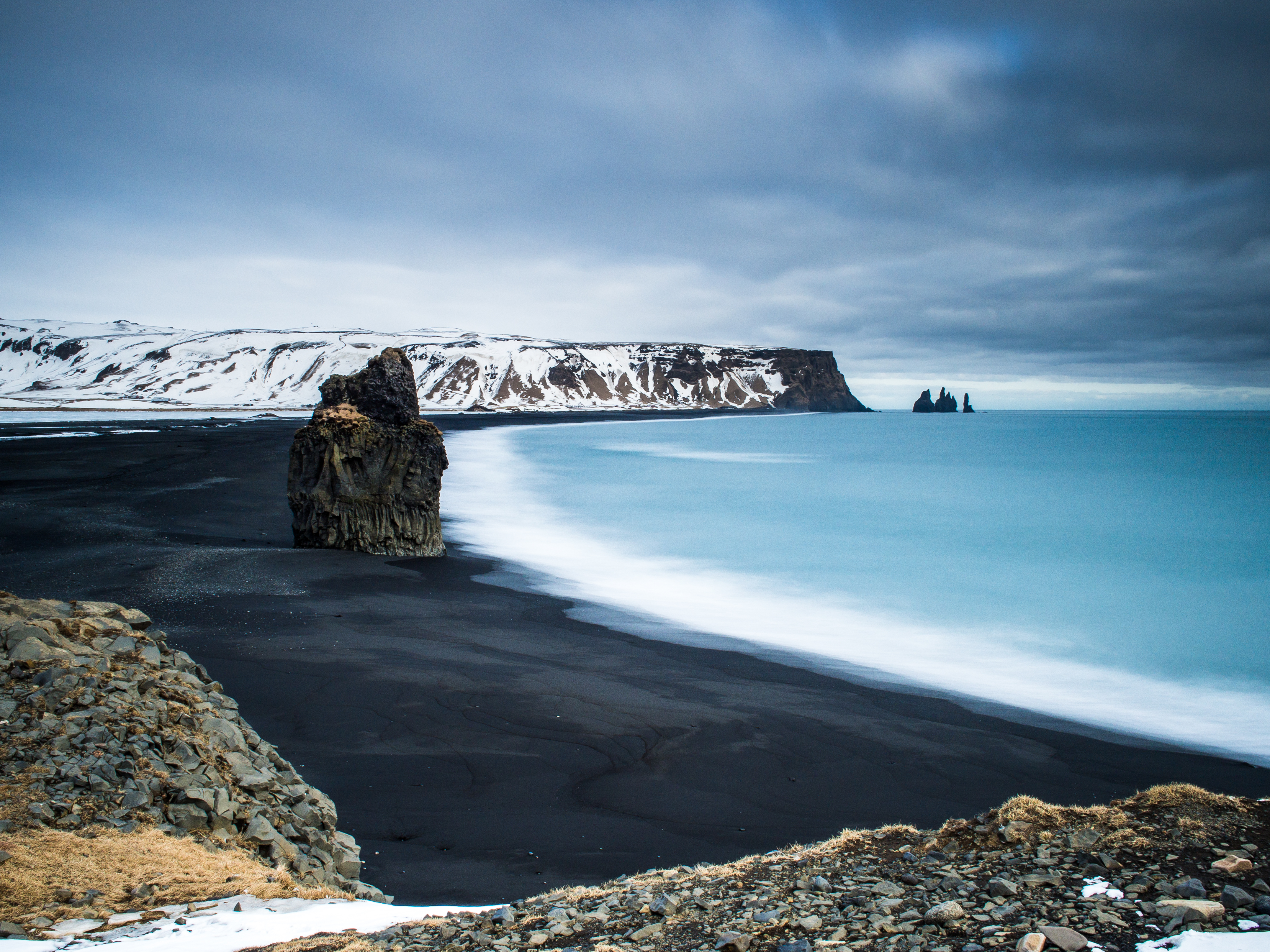 Schwarzer Sandstrand mit Felsen und ruhigem Wasser in Vik, Island, unter bewölktem Himmel.