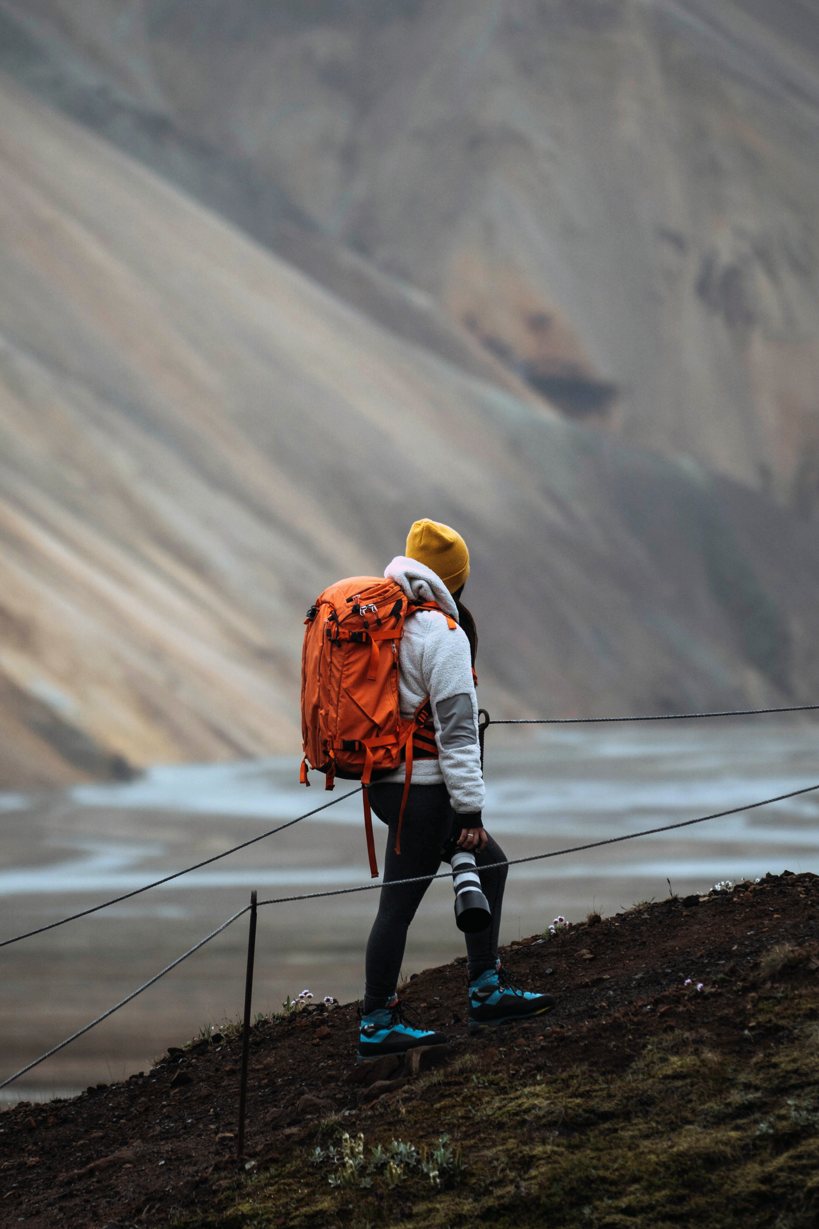 Person mit orangefarbenem Rucksack und Kamera, die in einer bergigen Landschaft steht.