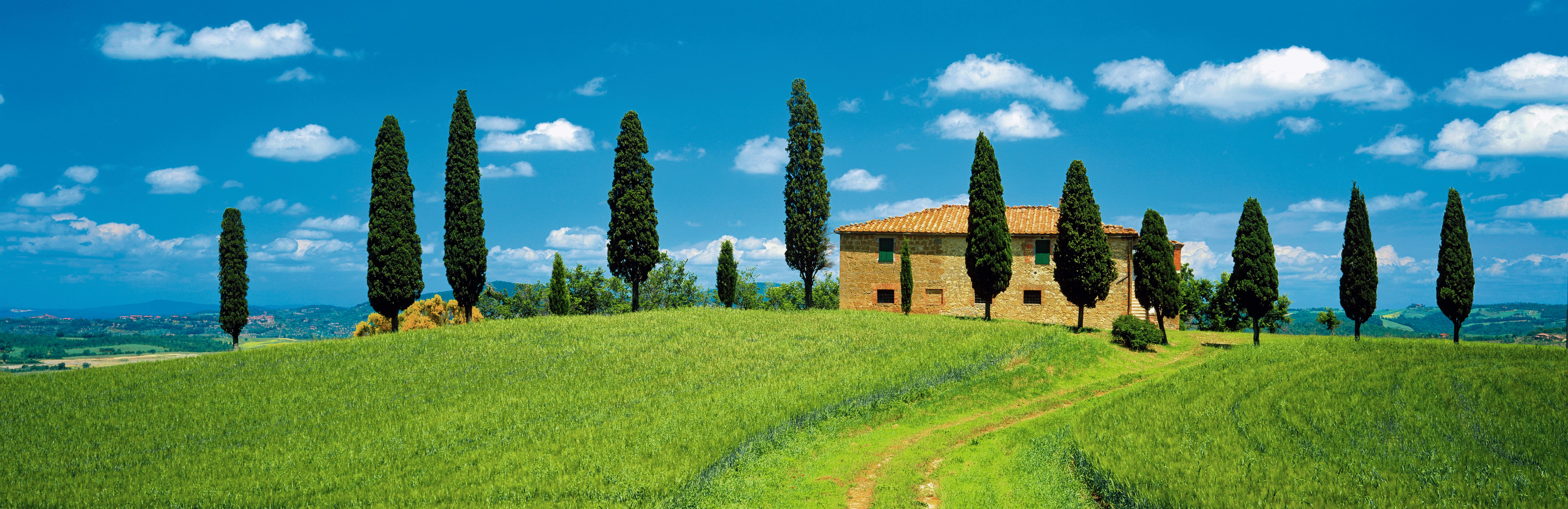 Landschaft mit einem traditionellen toskanischen Bauernhaus, umgeben von Zypressen und grünen Feldern unter blauem Himmel.
