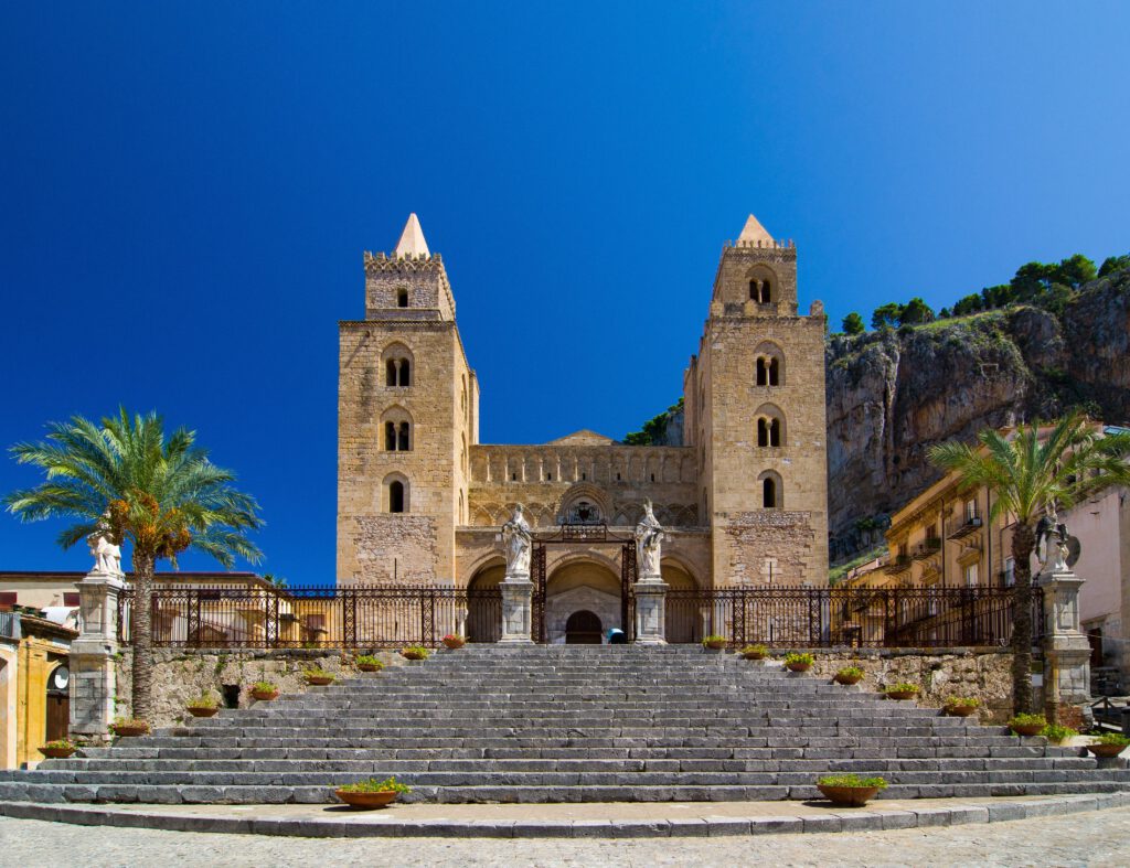 Normannendom in Cefalù mit zwei Türmen und einer Treppe, umgeben von Palmen und blauem Himmel.