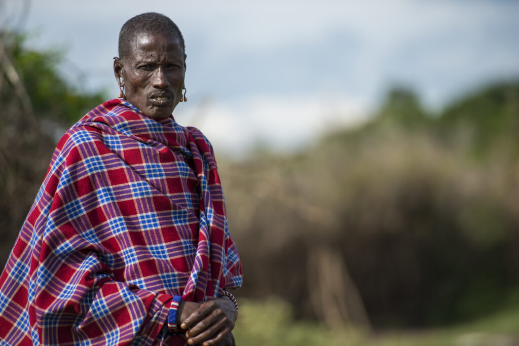 Ein Maasai-Mann in traditioneller Kleidung mit einem rot-blauen karierten Shuka, steht im Freien.