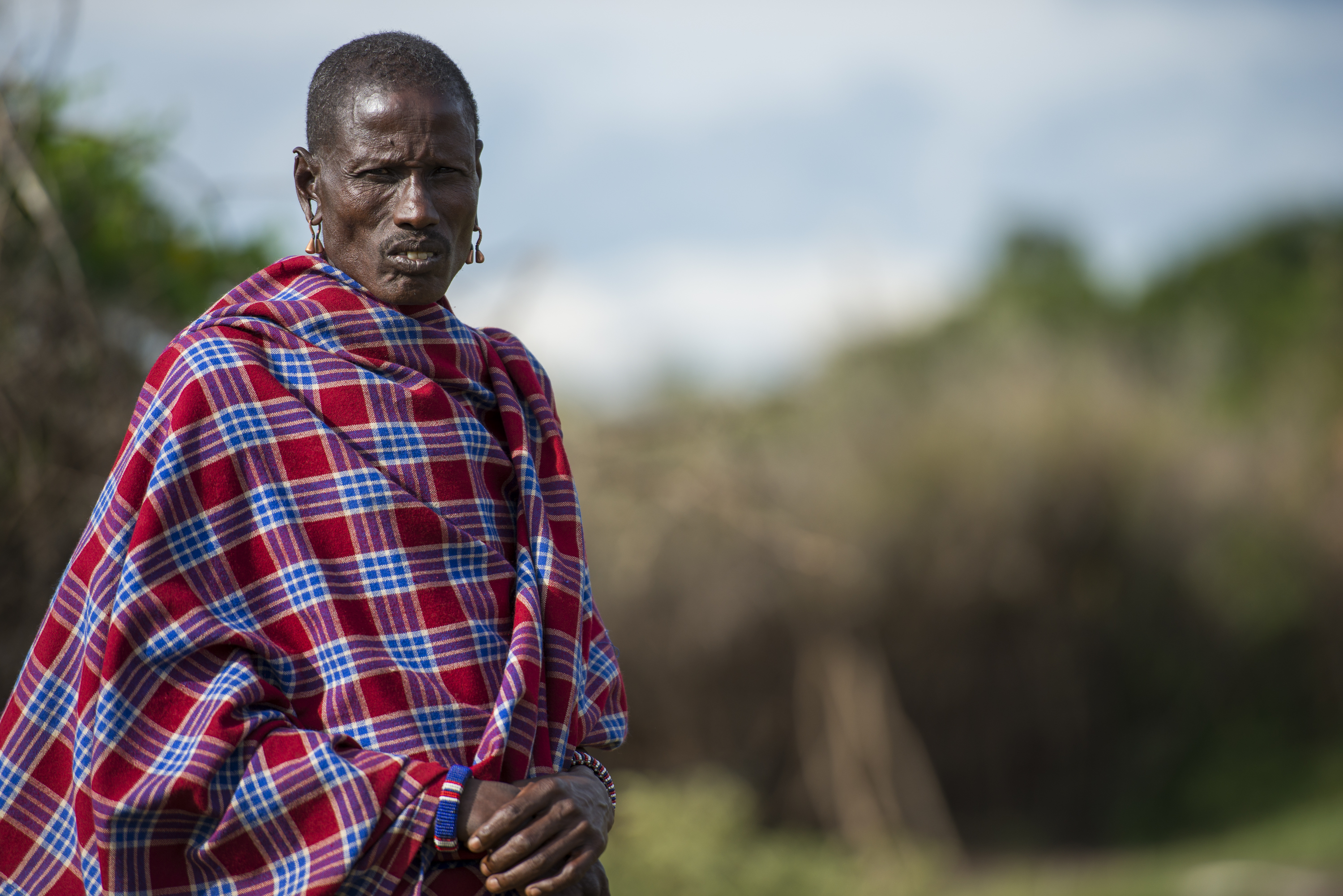 Ein Maasai-Mann in traditioneller Kleidung mit einem rot-blauen karierten Shuka, steht im Freien.
