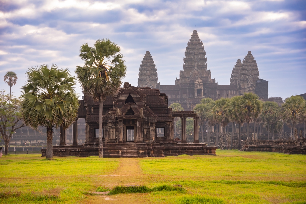 Angkor Wat Tempelkomplex in Kambodscha mit Palmen im Vordergrund und Wolken am Himmel.