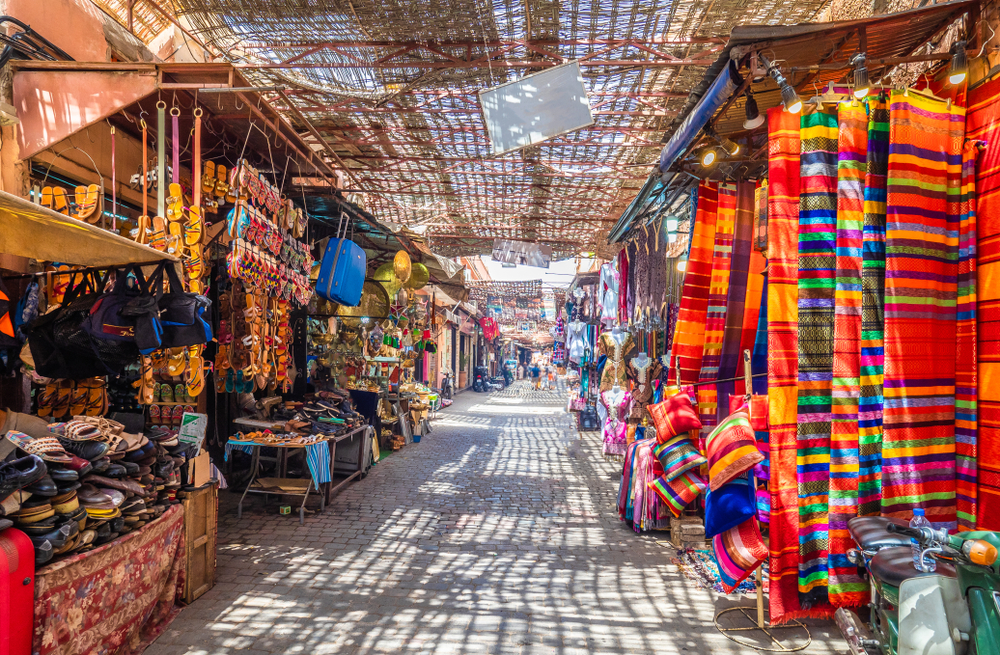 Bunte Stoffe und Waren in einem Souk in Marrakesch, mit Schatten von einem Dach über den Ständen.