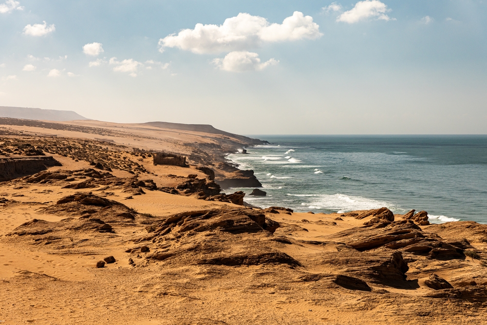 Küstenlandschaft mit Sanddünen und Wellen, die gegen die Felsen schlagen.