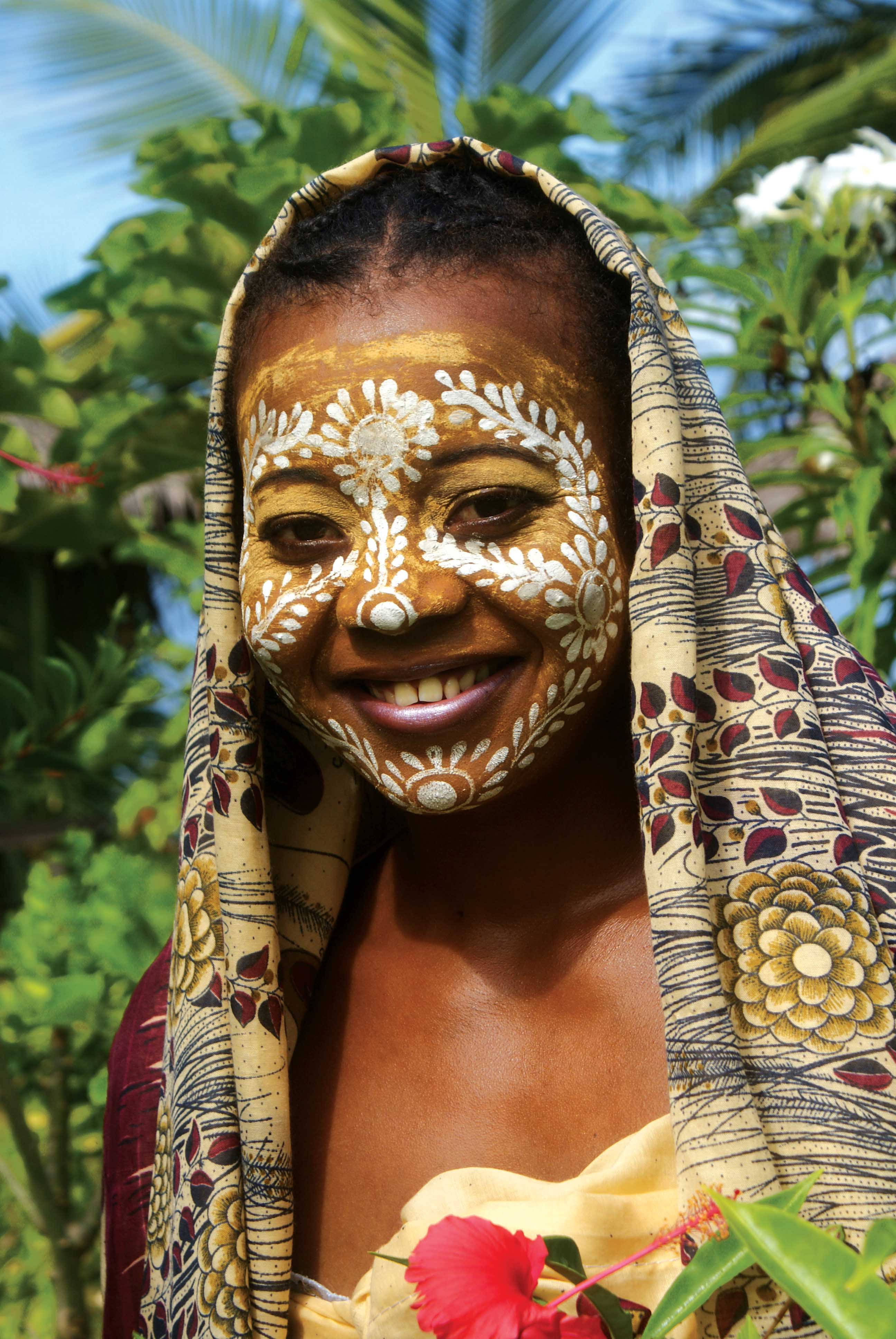 Frau mit traditioneller Gesichtsbemalung und Kopftuch, umgeben von Pflanzen und Blumen.