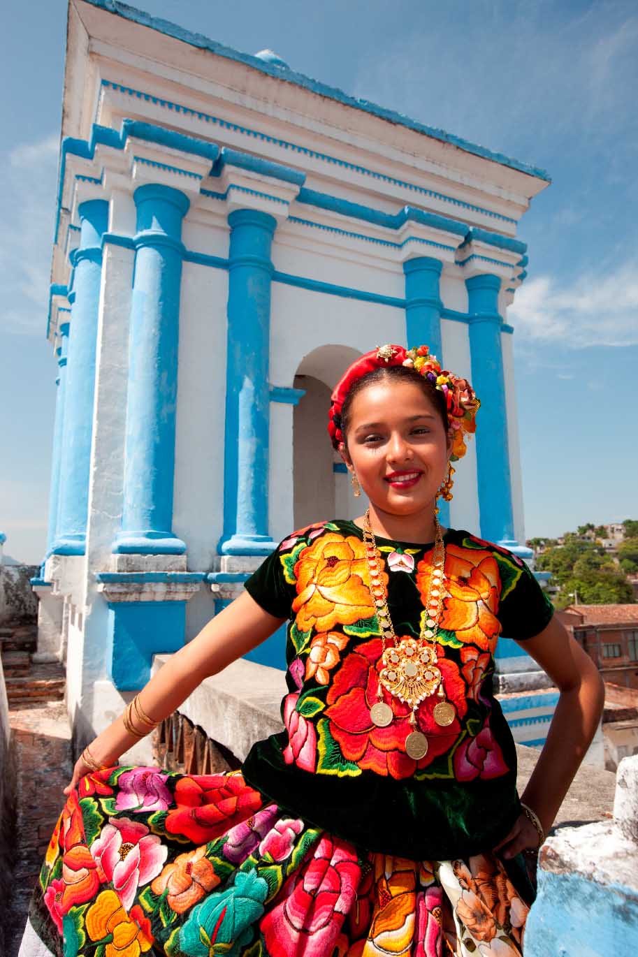Junge Frau in traditioneller mexikanischer Tracht mit bunten Blumenmustern und Schmuck auf einem Dach.