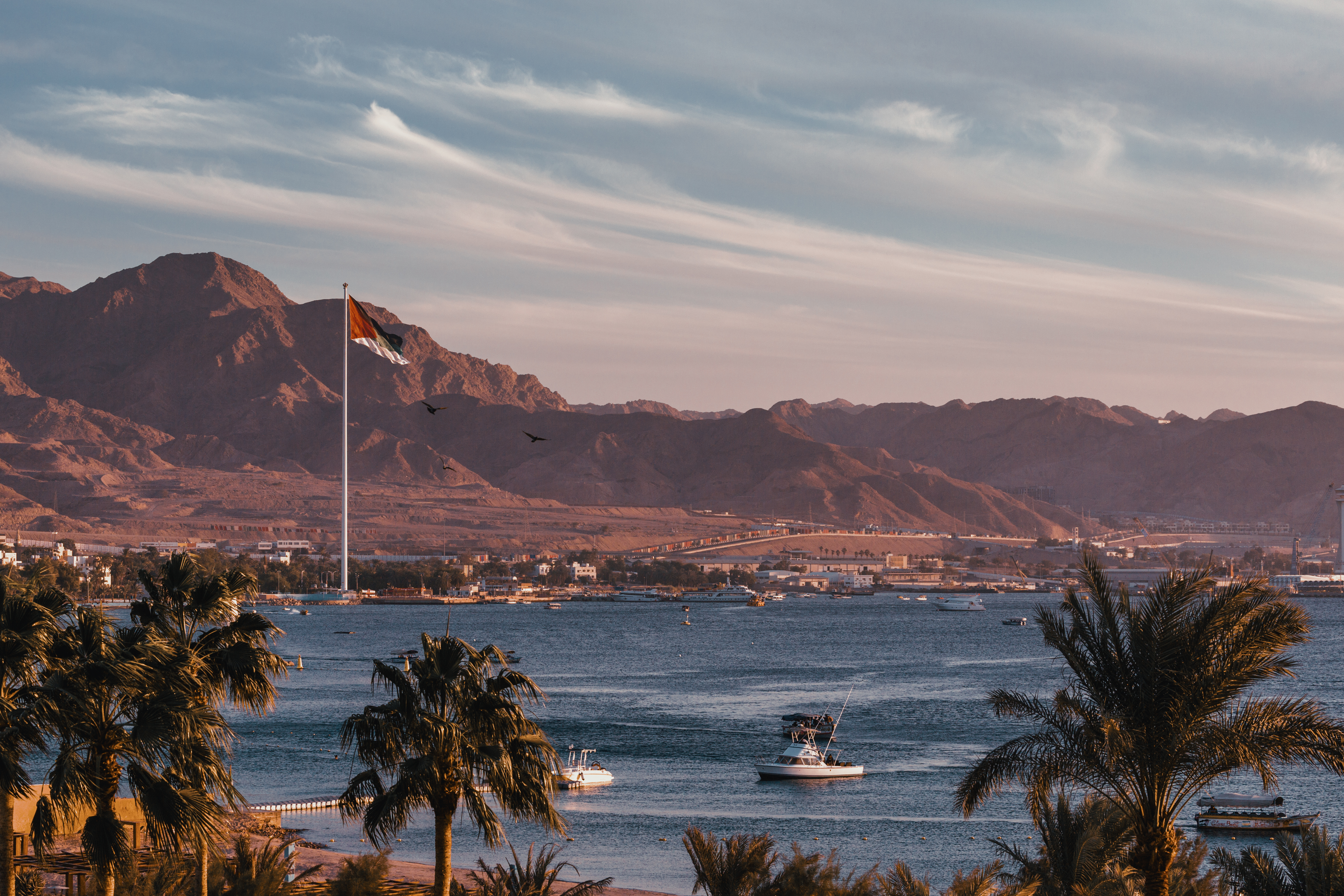Blick auf das Wasser mit Booten und einer großen Flagge vor einer Berglandschaft bei Sonnenuntergang.