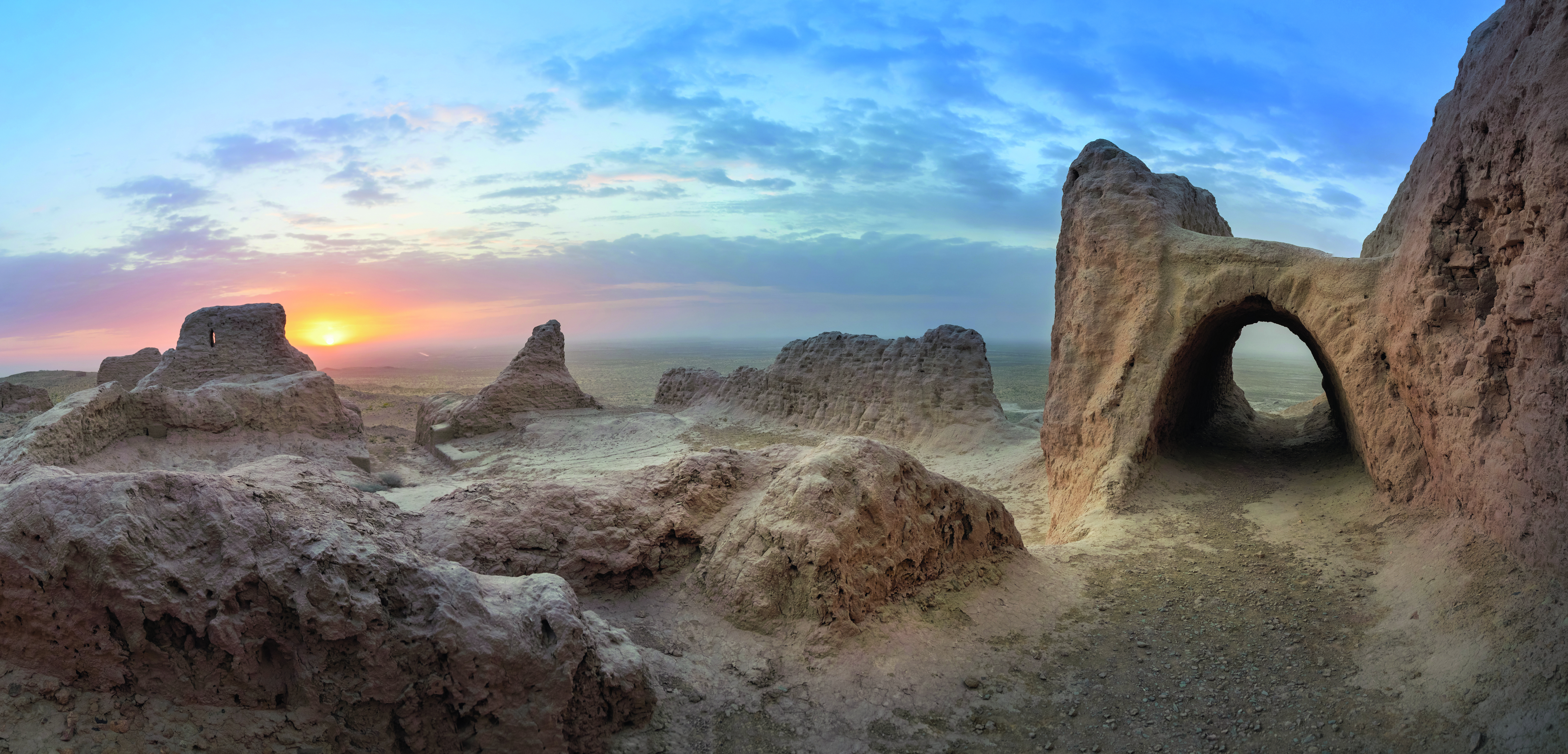 Panoramaaufnahme einer alten Ruine mit Felsen und einem Bogen, der in den Himmel ragt, bei Sonnenuntergang.