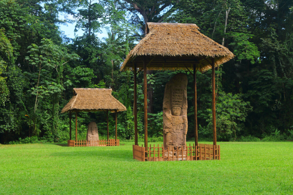 Zwei steinerne Monumente unter Strohdächern in einer grünen Wiese umgeben von Bäumen.