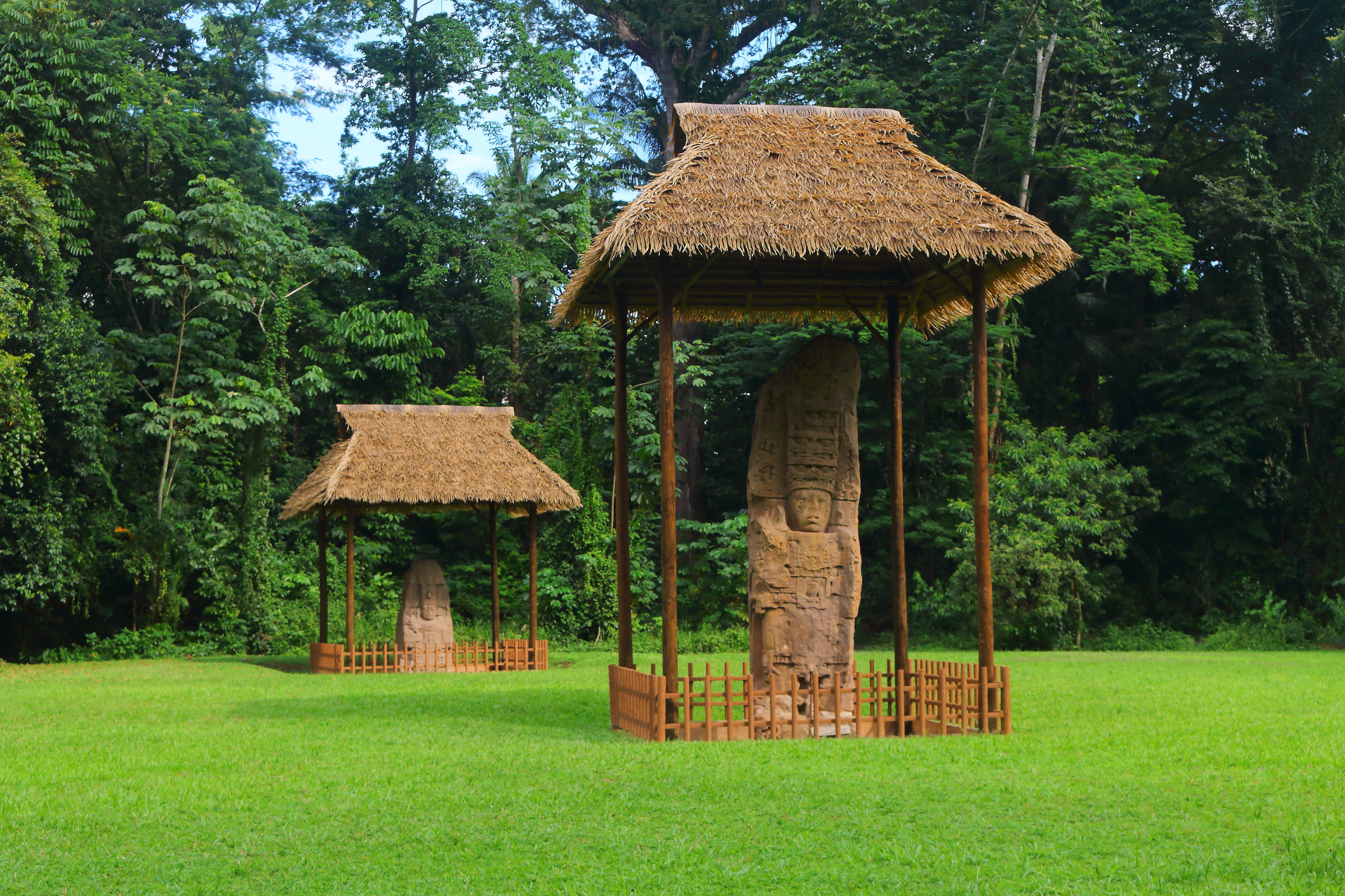 Zwei steinerne Monumente unter Strohdächern in einer grünen Wiese umgeben von Bäumen.