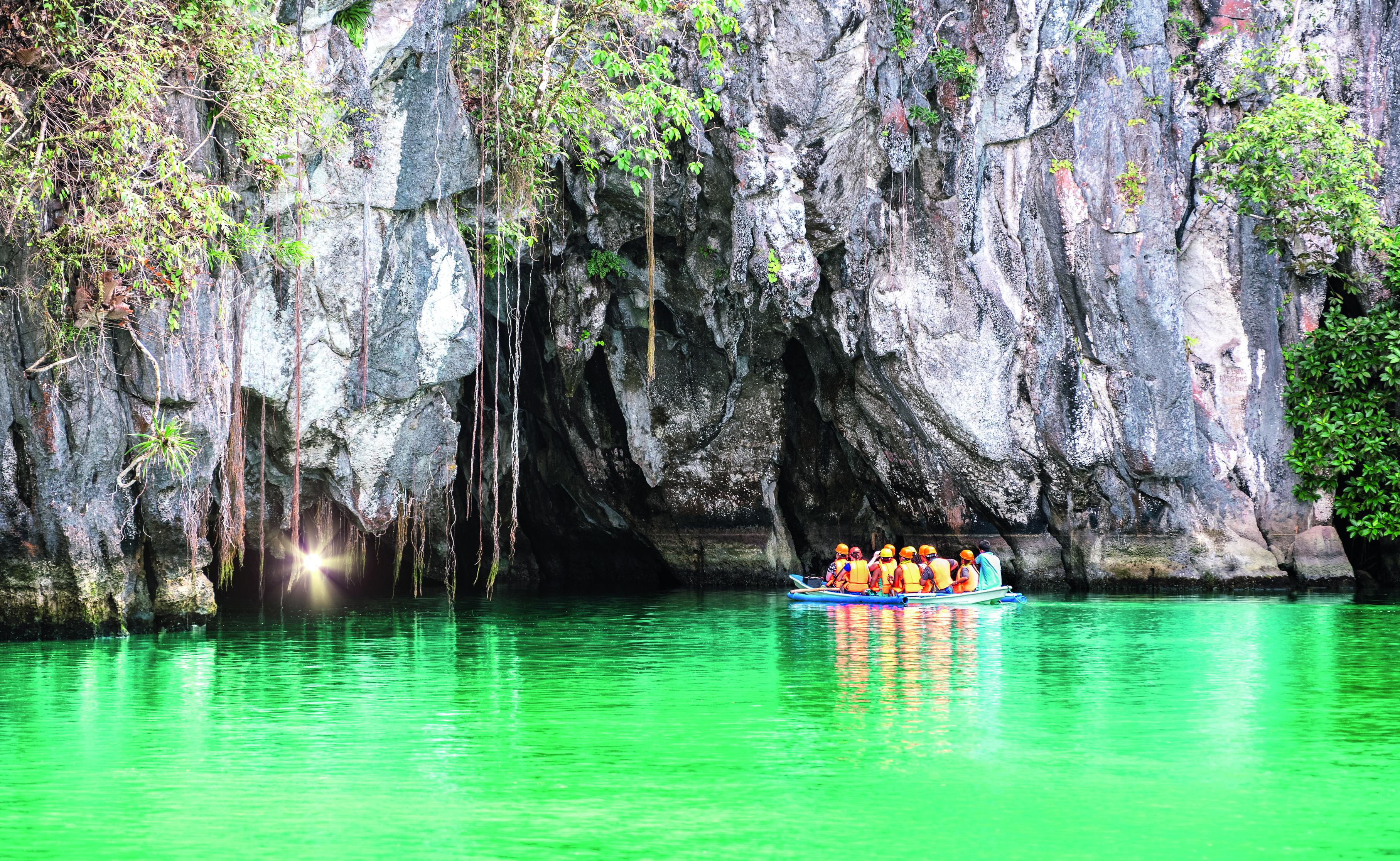 Gruppe von Personen in einem Boot auf grünem Wasser vor einer Felsformation mit Pflanzen und einem Höhleneingang.