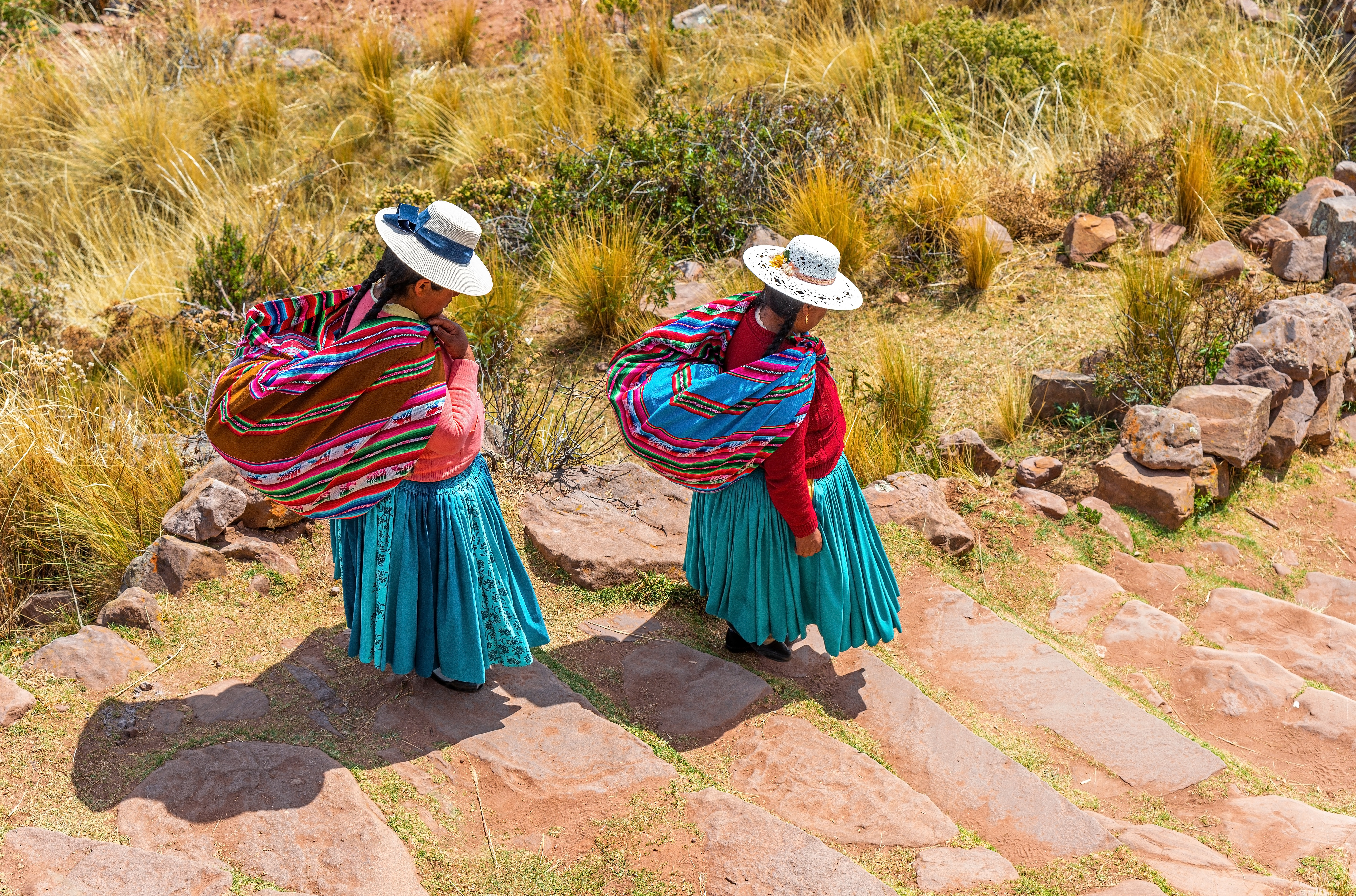 Zwei Frauen in traditioneller peruanischer Tracht mit bunten Umhängen und Hüten auf einem Steinweg.