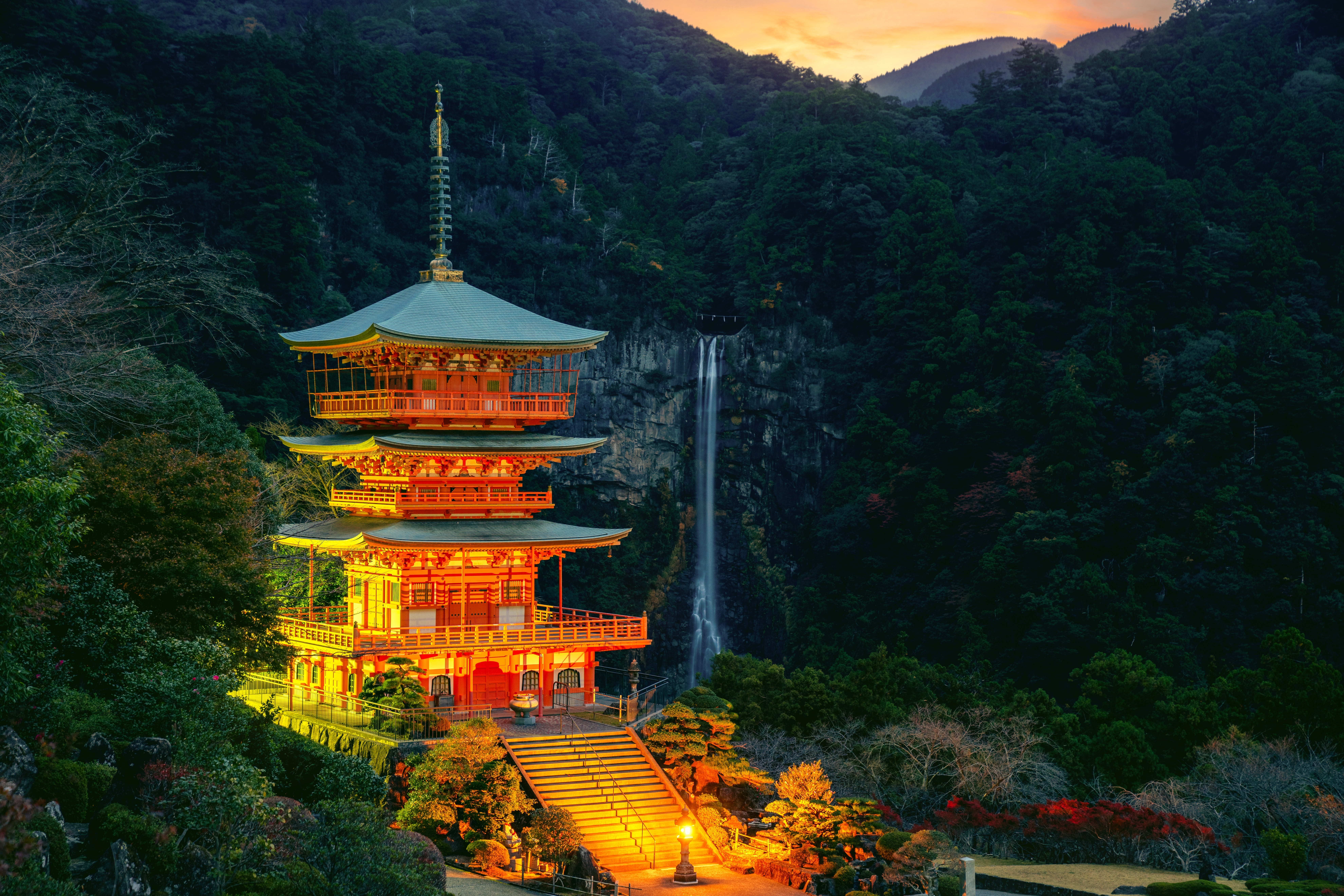 Pagode mit leuchtenden Farben und Wasserfall im Hintergrund, umgeben von Bäumen und Bergen.