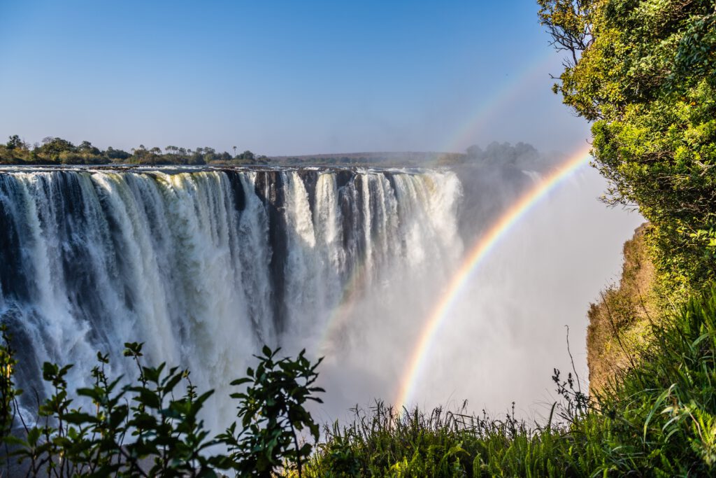 Victoriafälle mit einem Regenbogen, der über den Wasserfall gespannt ist, umgeben von üppigem Grün.