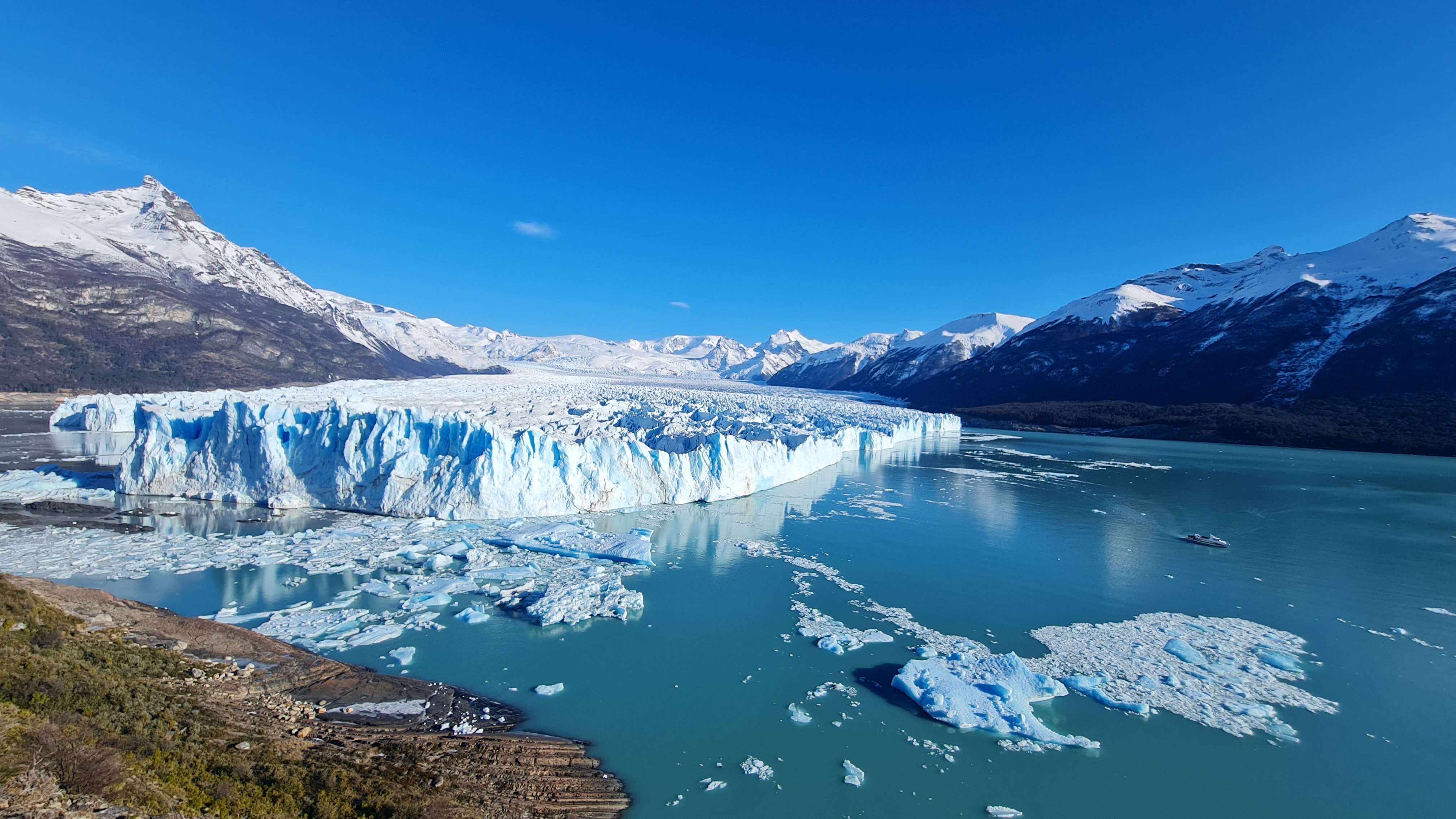 Gletscher mit blauen Eisformationen und schneebedeckten Bergen im Hintergrund unter klarem Himmel.