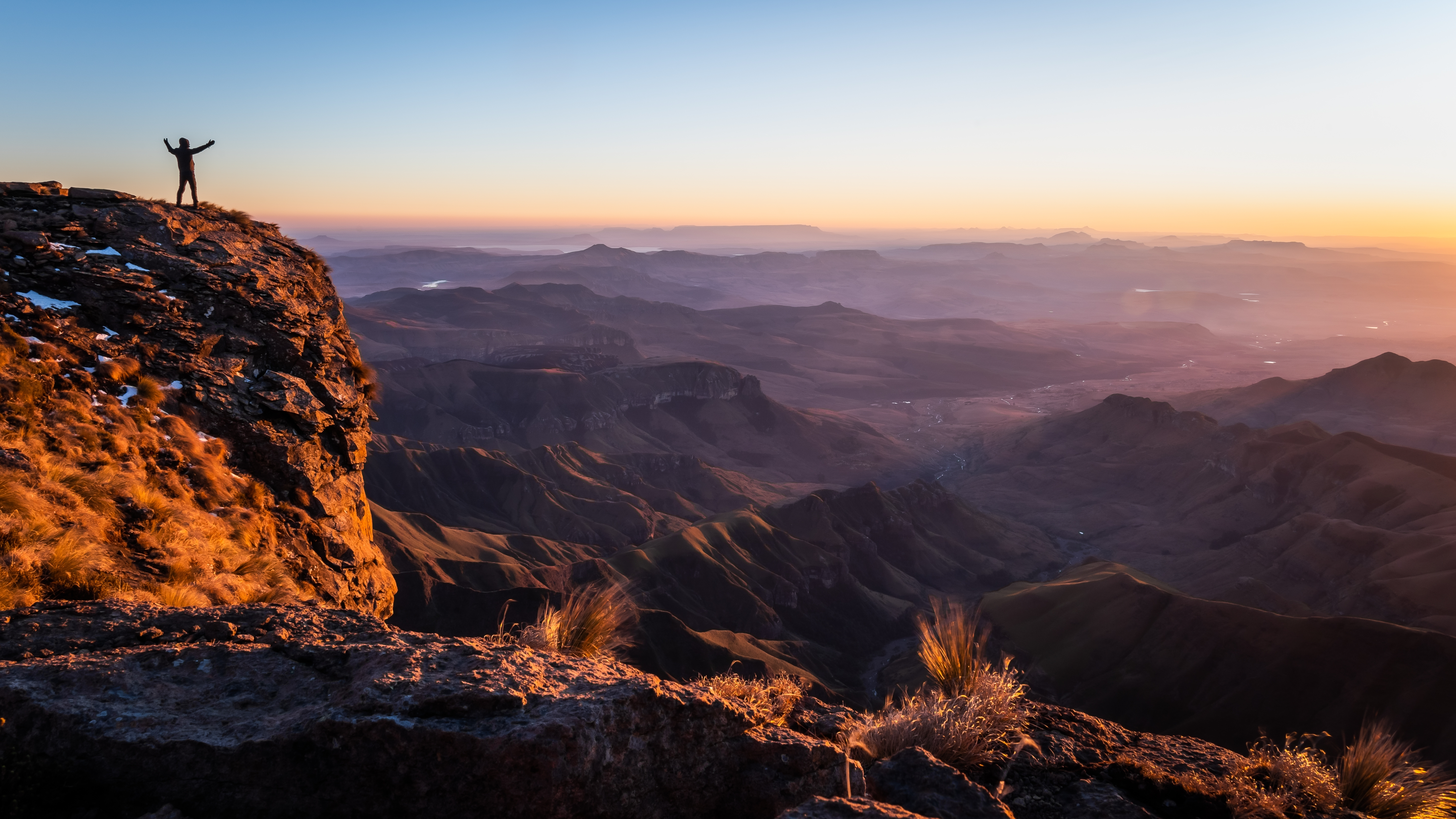 Person steht auf einem Felsen und blickt auf eine weite Berglandschaft bei Sonnenuntergang.