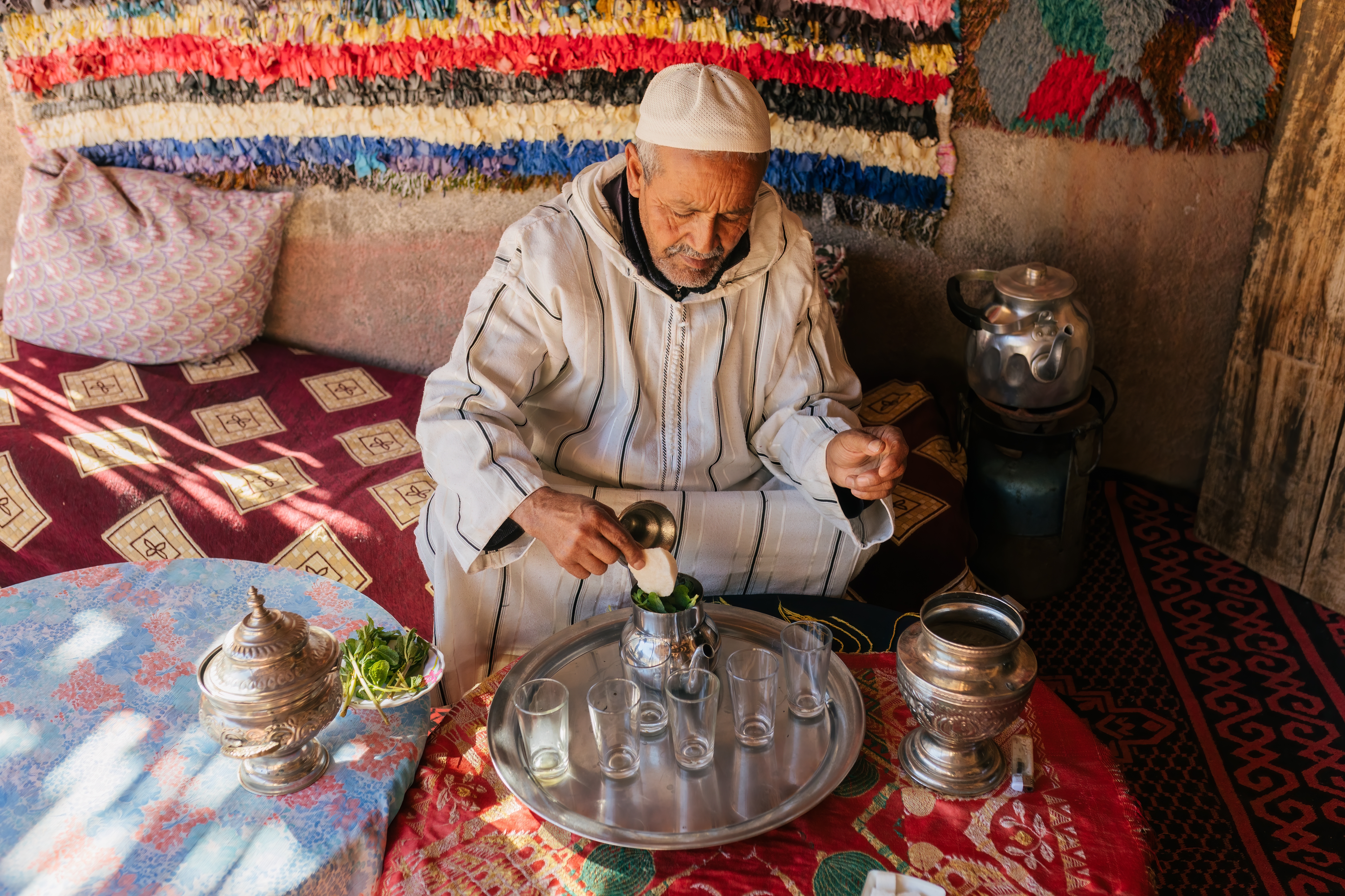 Ein älterer Mann in traditioneller Kleidung bereitet Tee mit frischen Kräutern in einem Raum mit bunten Textilien.