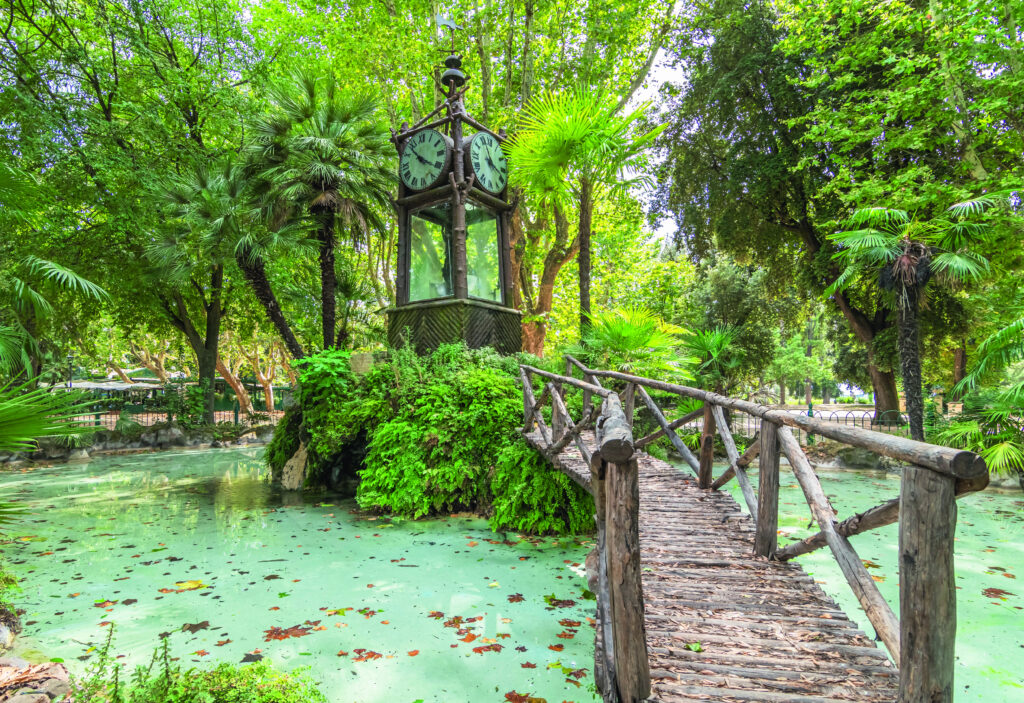 Eine alte Uhr mit Ziffernblatt und Zeigern, umgeben von üppigem Grün und einem Holzsteg über einen Teich.