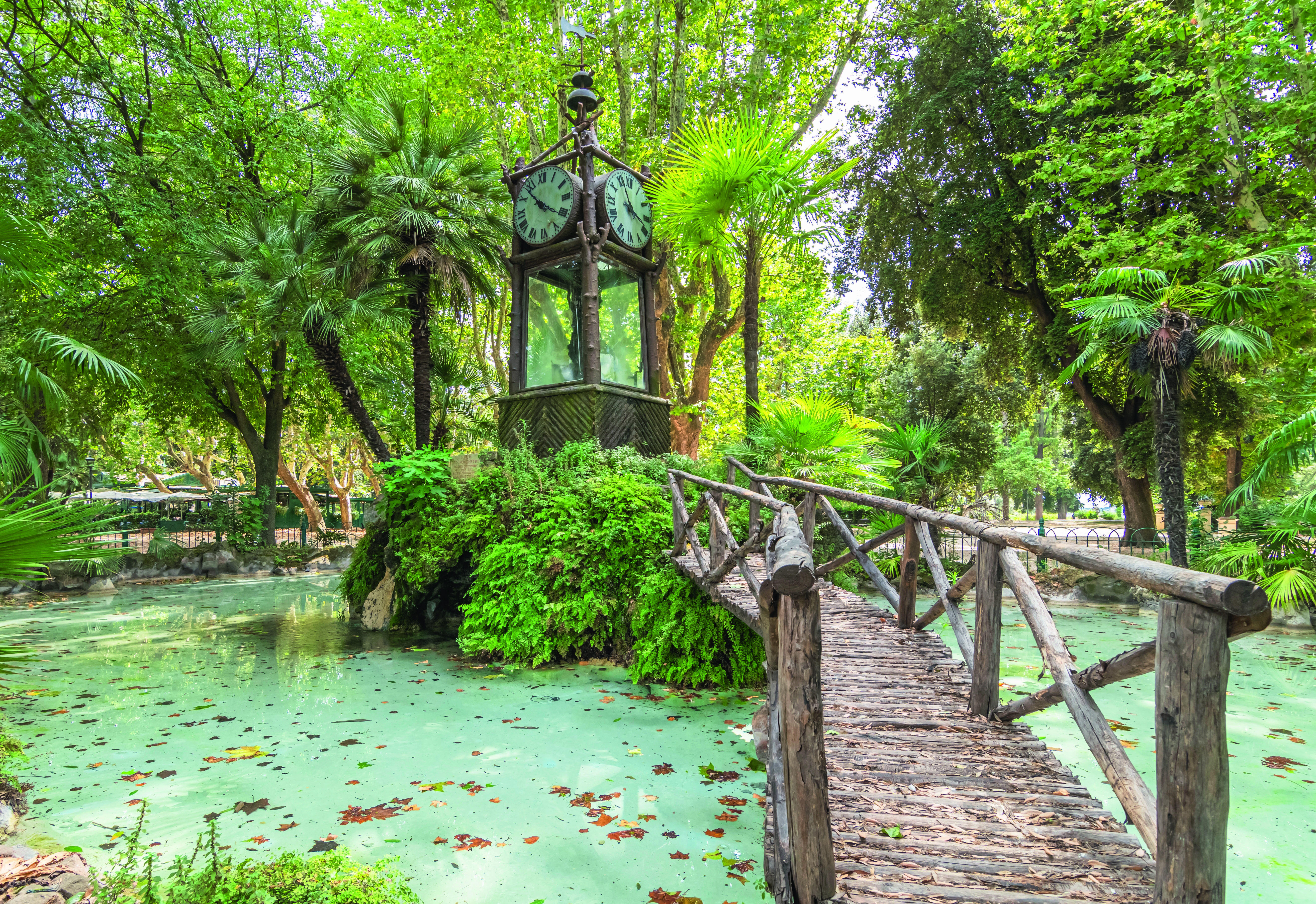 Eine alte Uhr mit Ziffernblatt und Zeigern, umgeben von üppigem Grün und einem Holzsteg über einen Teich.