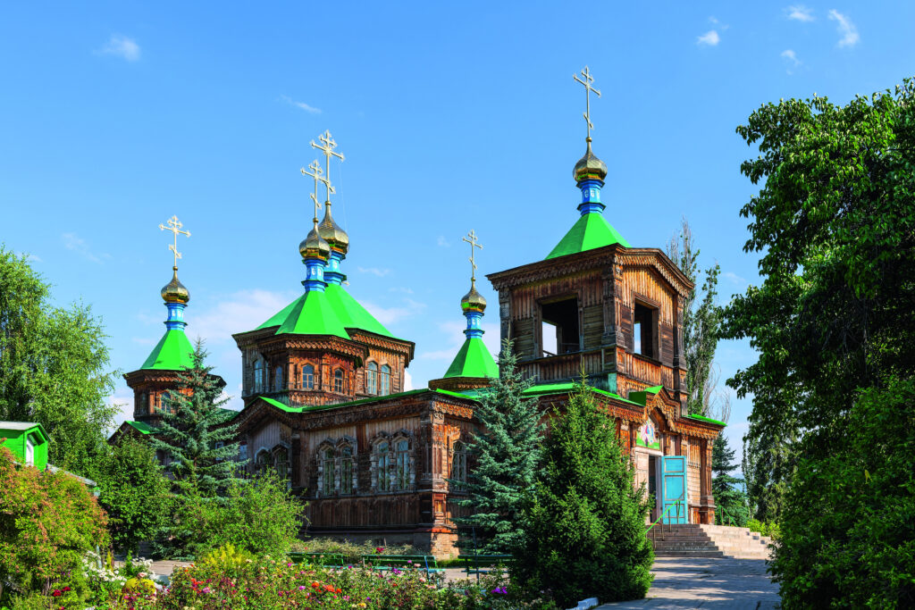 Holzkirche mit grünen Dächern und goldenen Kuppeln, umgeben von Bäumen und Blumen.
