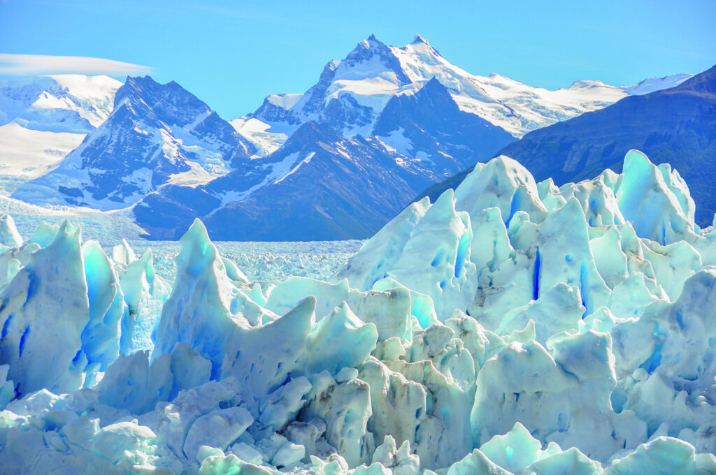 Eisbergformationen mit blauen und weißen Farbtönen vor schneebedeckten Bergen im Hintergrund.
