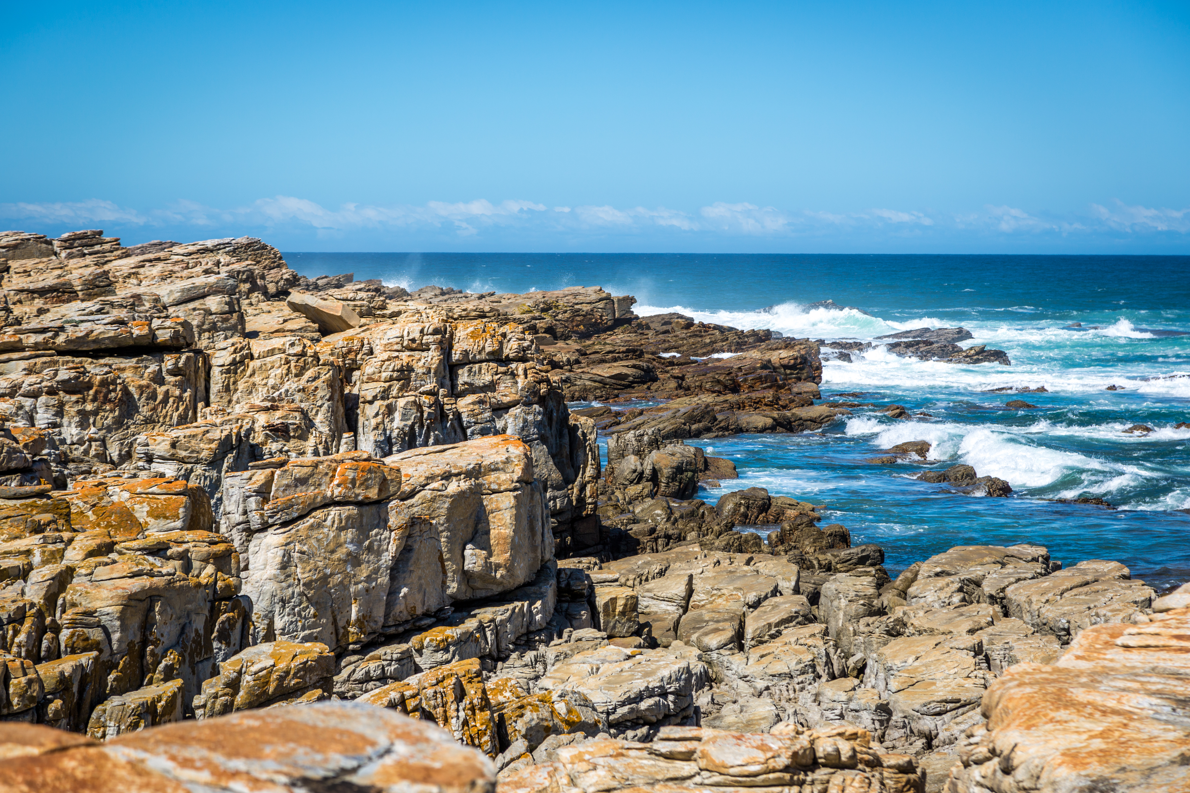 Felsen und Küstenlinie mit Wellen, die gegen die Klippen schlagen, unter klarem blauen Himmel.