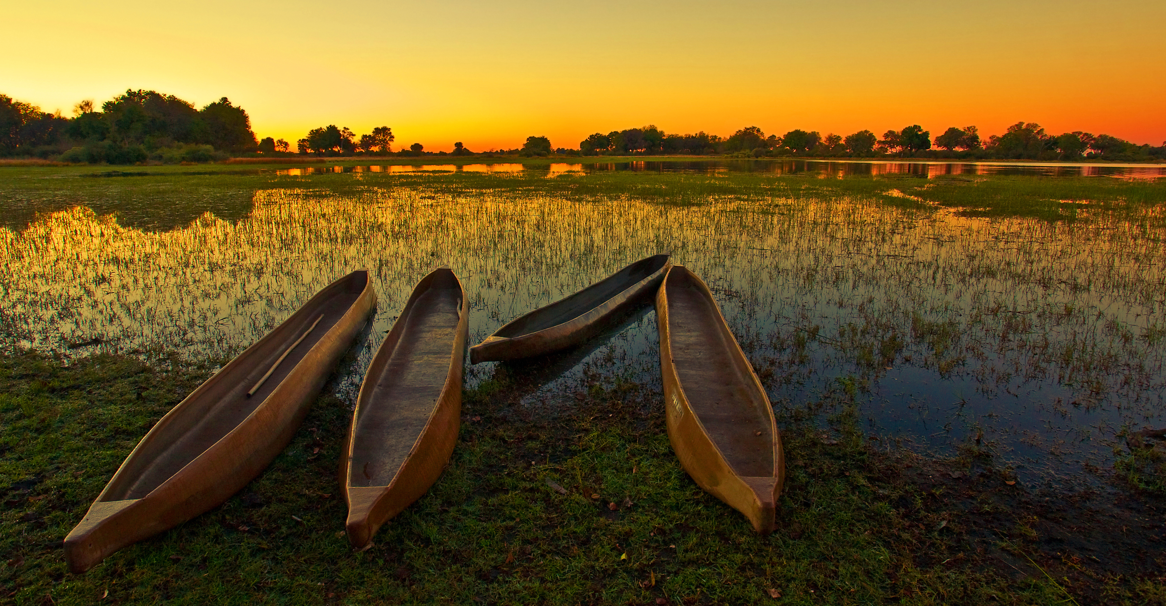 Vier traditionelle Holzkanus am Ufer eines ruhigen Gewässers bei Sonnenuntergang.