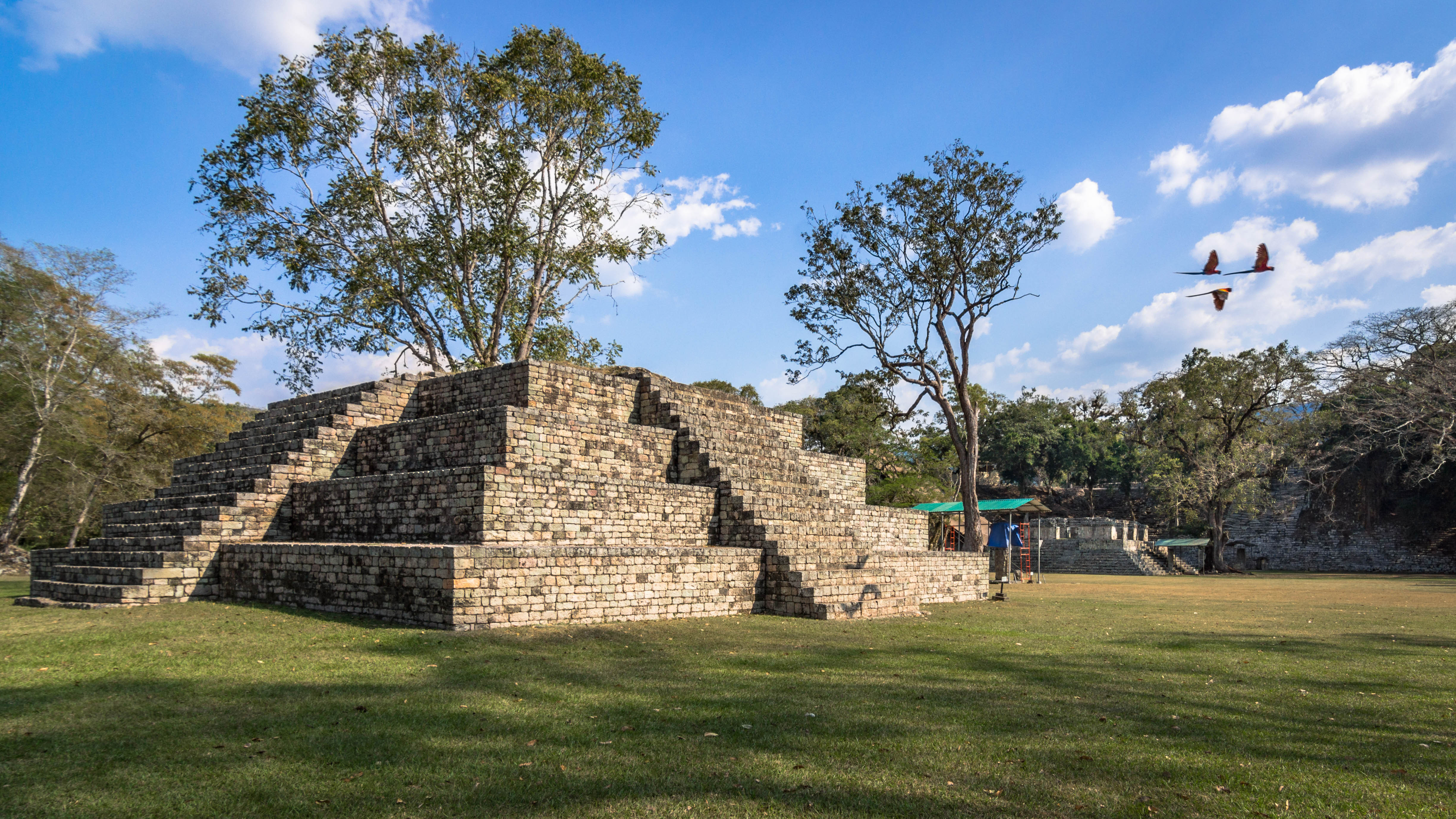 Steinerne Pyramide mit Stufen und Bäumen im Hintergrund, unter blauem Himmel mit Wolken.