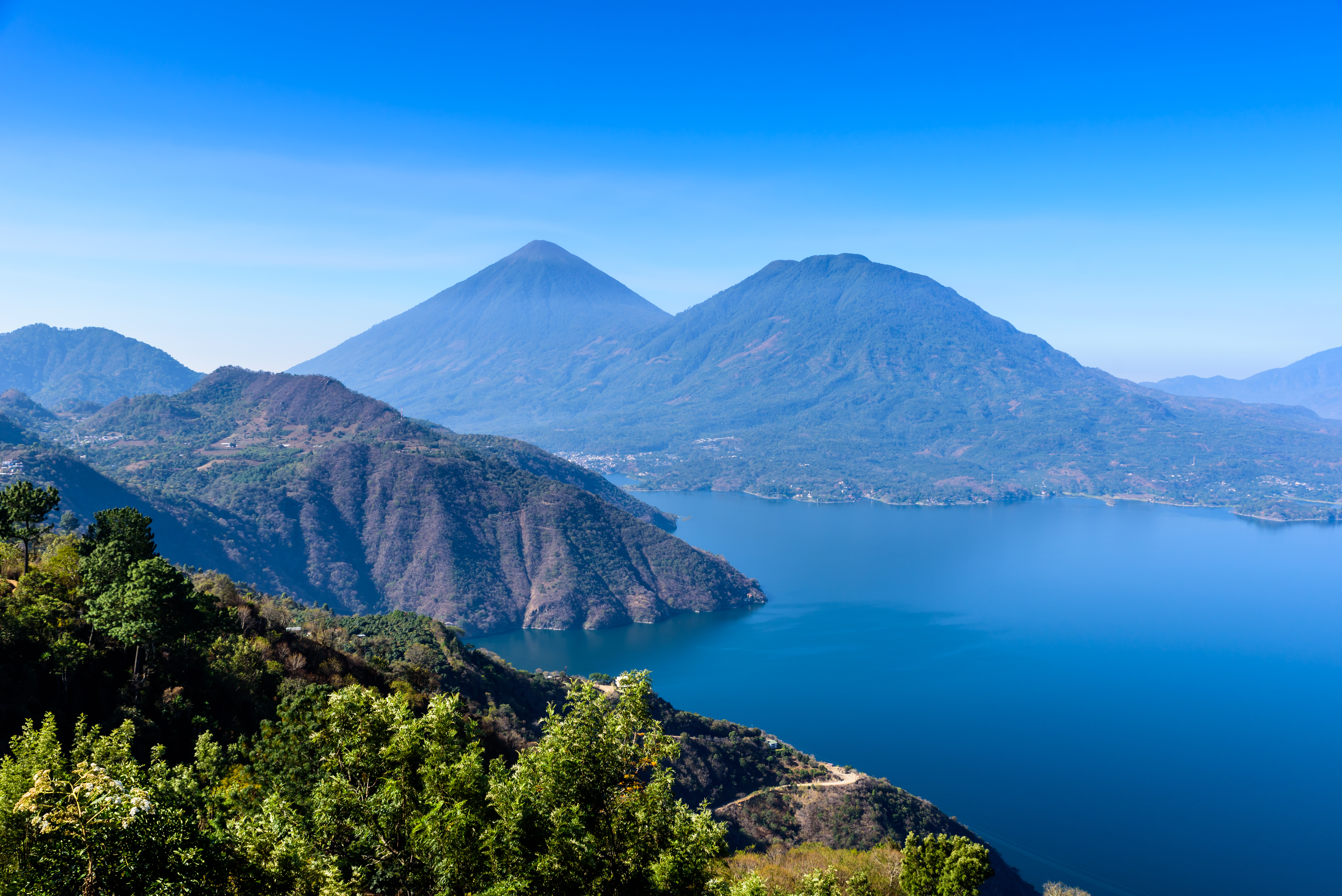 Blick auf den Atitlán-See mit Vulkanen im Hintergrund und klarem Himmel.